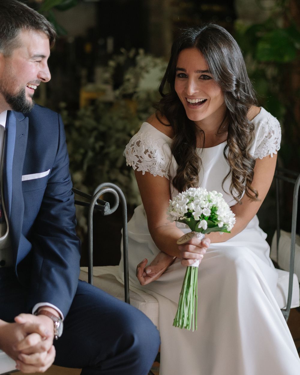 A bride in a white lace gown smiles while holding a small bouquet next to her groom in a navy suit.