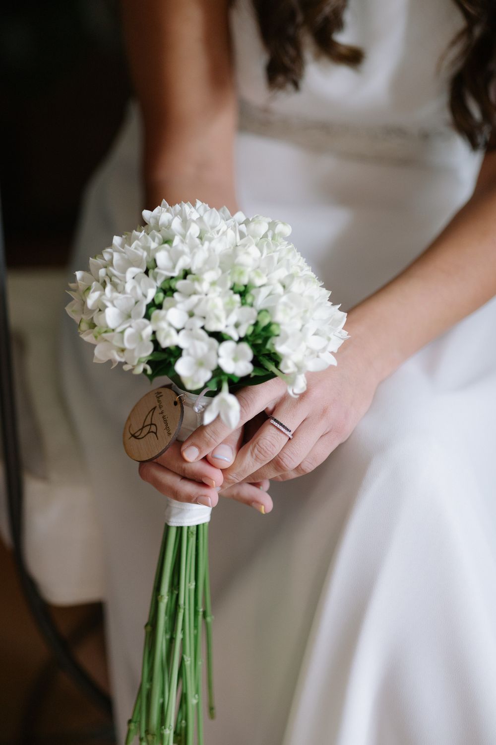 A wedding bouquet of white freesias wrapped in twine held by hands wearing a simple gold band against a white dress.