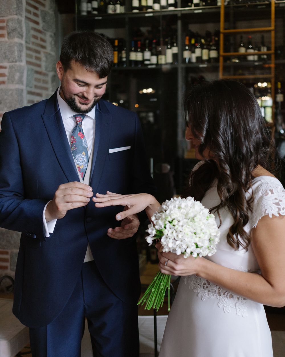 A couple exchanges vows in a wine cellar-style venue with white floral bouquet and navy suit attire.