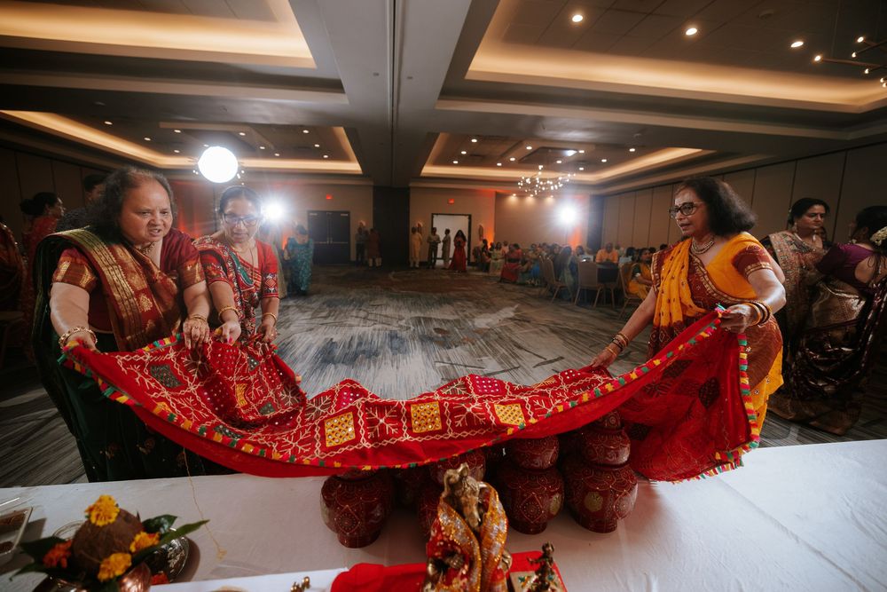Traditional Indian wedding ceremony with people holding a long red and gold ceremonial garment in an elegant banquet hall.