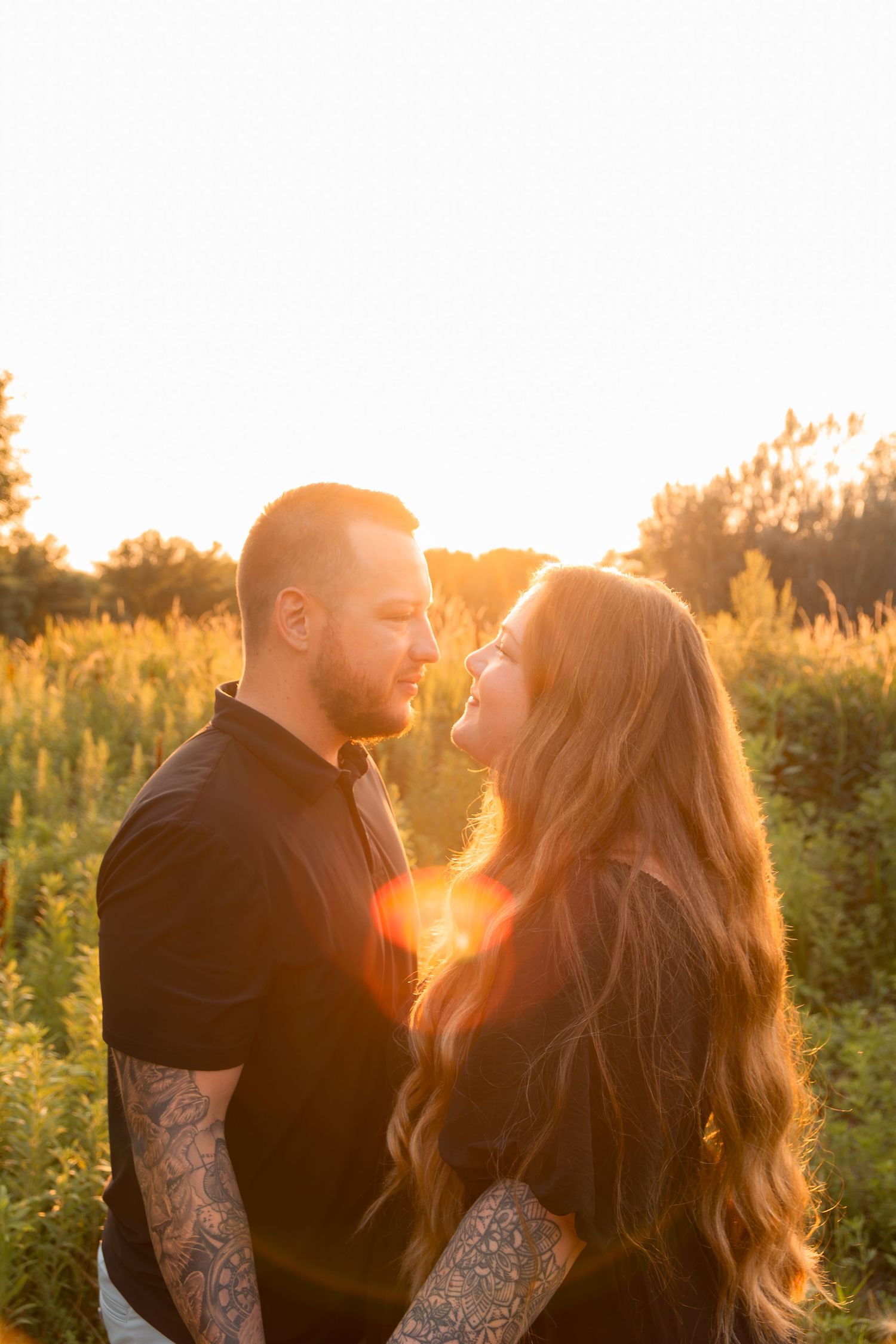 Couple facing each other in golden sunset light in a field, man with tattoos in black shirt, woman with long hair.