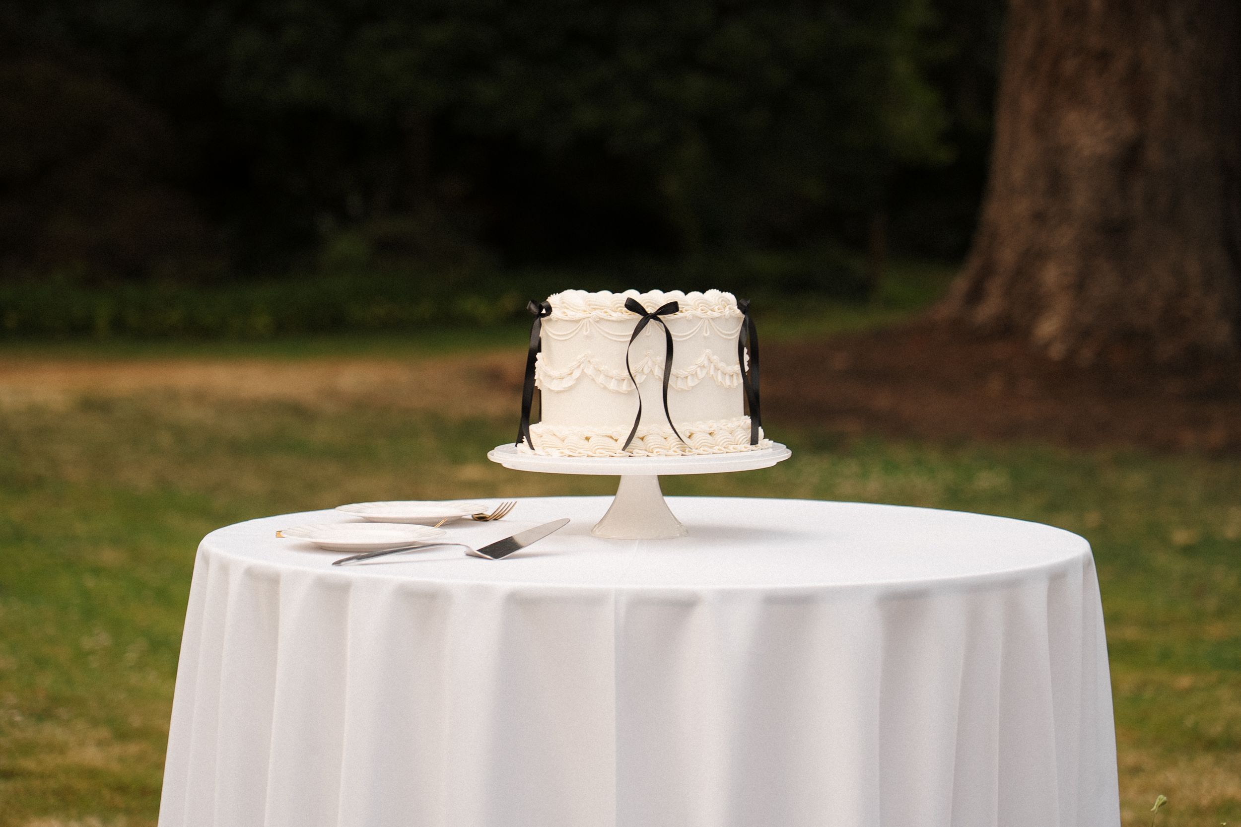 Small white wedding cake with black ribbon detail sits atop round table covered in white linen outdoors.