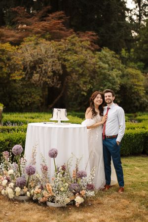 Romantic outdoor sweetheart table with floral arrangements featuring white linens and purple allium blooms at sunset.