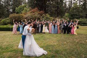 A newlywed couple shares a romantic moment while wedding guests cheer in a garden setting with evergreen trees.