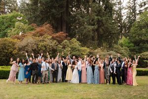 Wedding party celebrates outdoors with raised arms on a lawn surrounded by evergreen trees and autumn foliage.