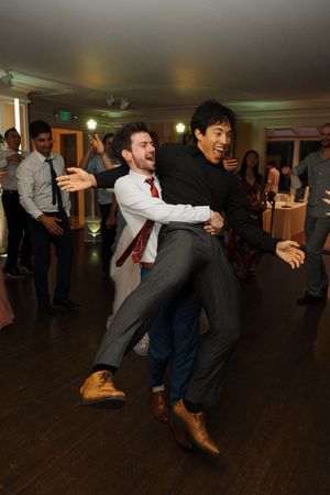 Party guests dancing in an energetic line formation on a dark wooden dance floor at an indoor celebration event.