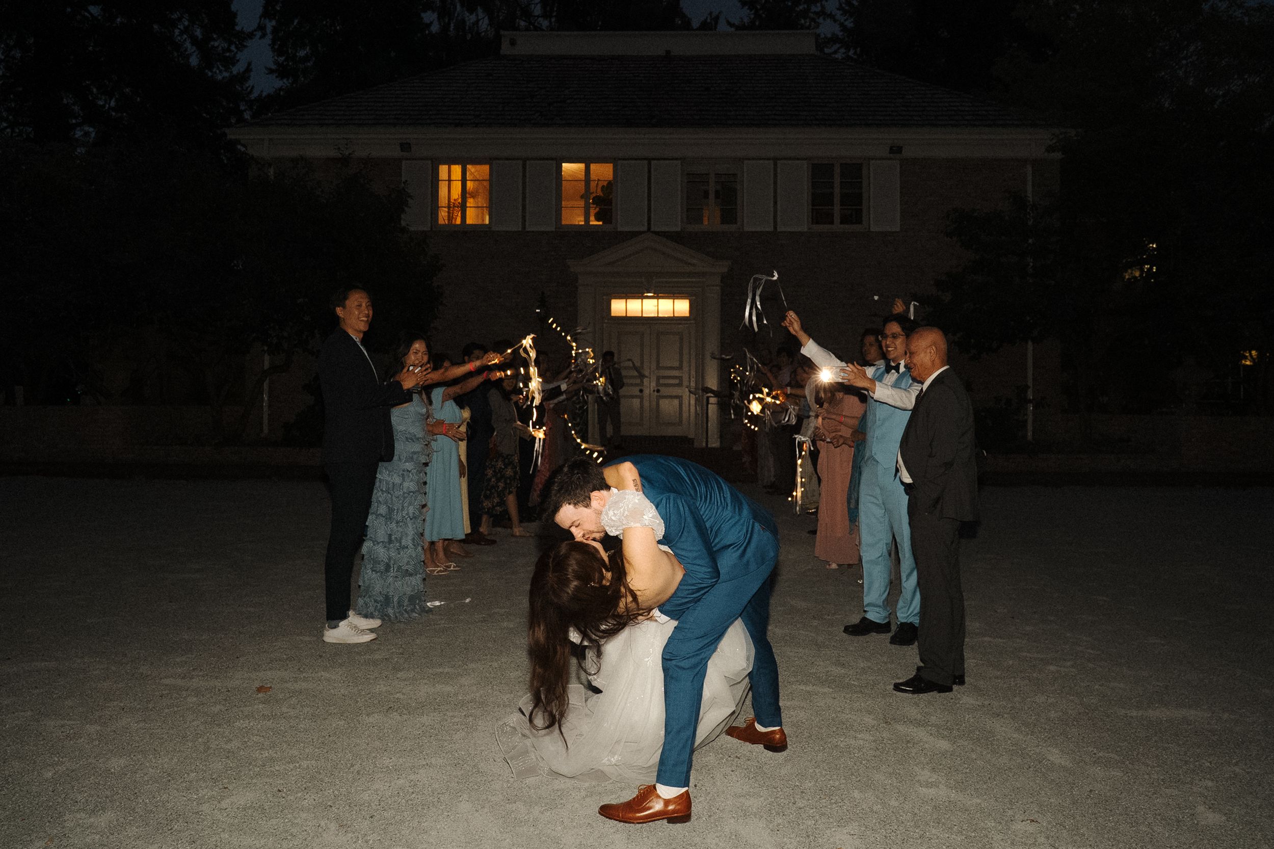 People dancing outdoors at night while others watch from a distance in a dimly lit area.