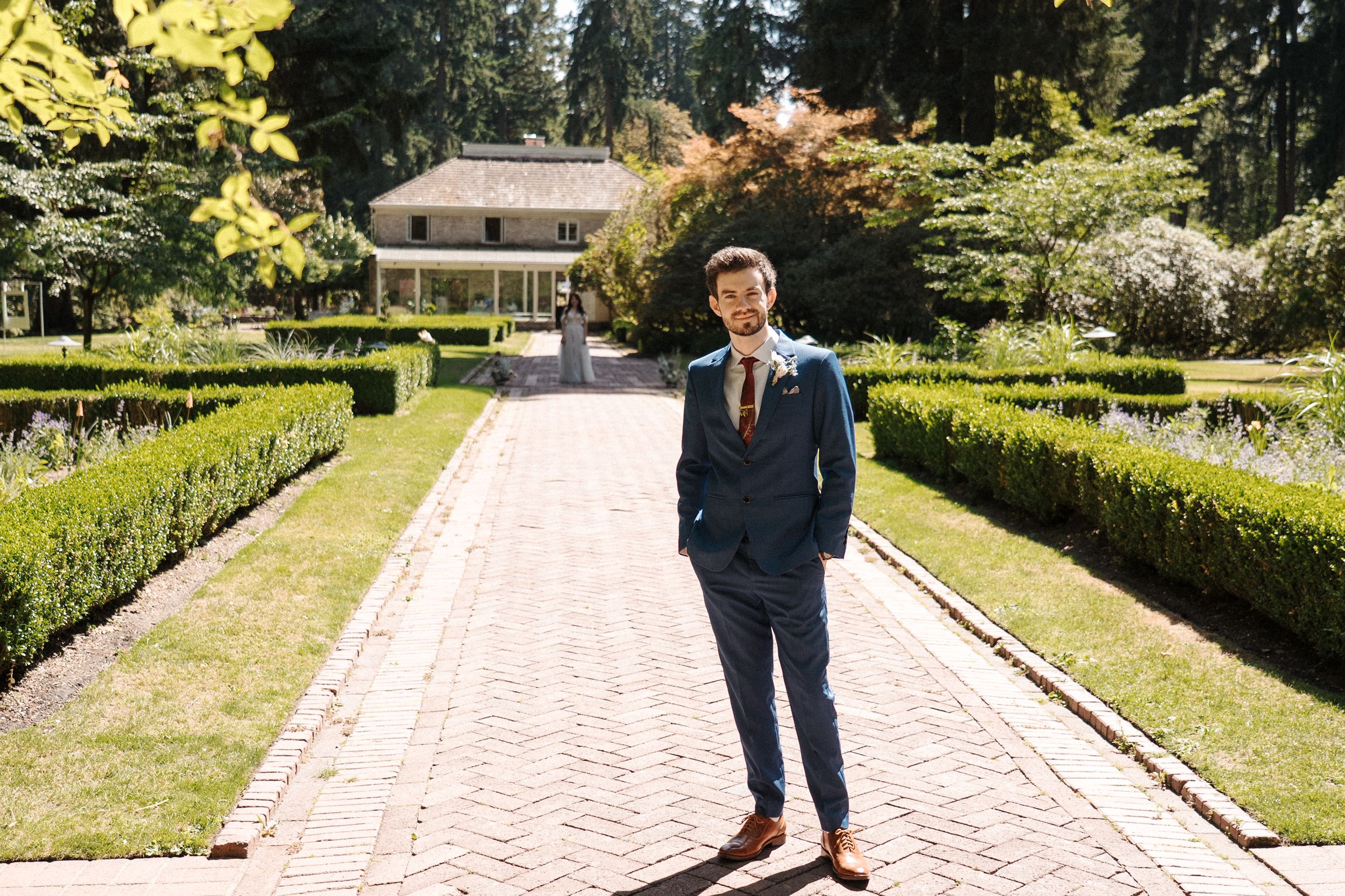 Well-dressed person in a navy suit stands on a brick pathway in a formal garden with a mansion in the background.