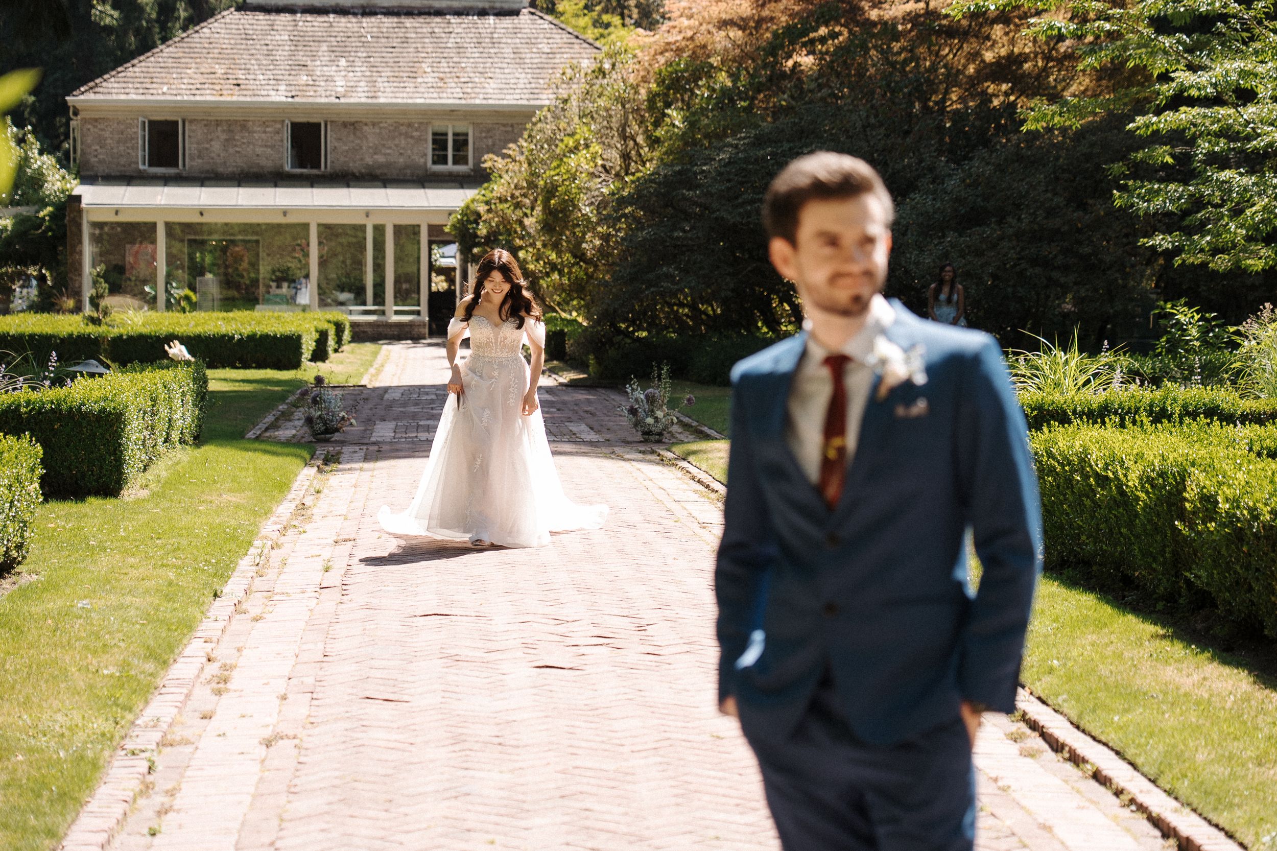 Wedding couple walks down stone garden path holding hands as sun shines through lush green trees.