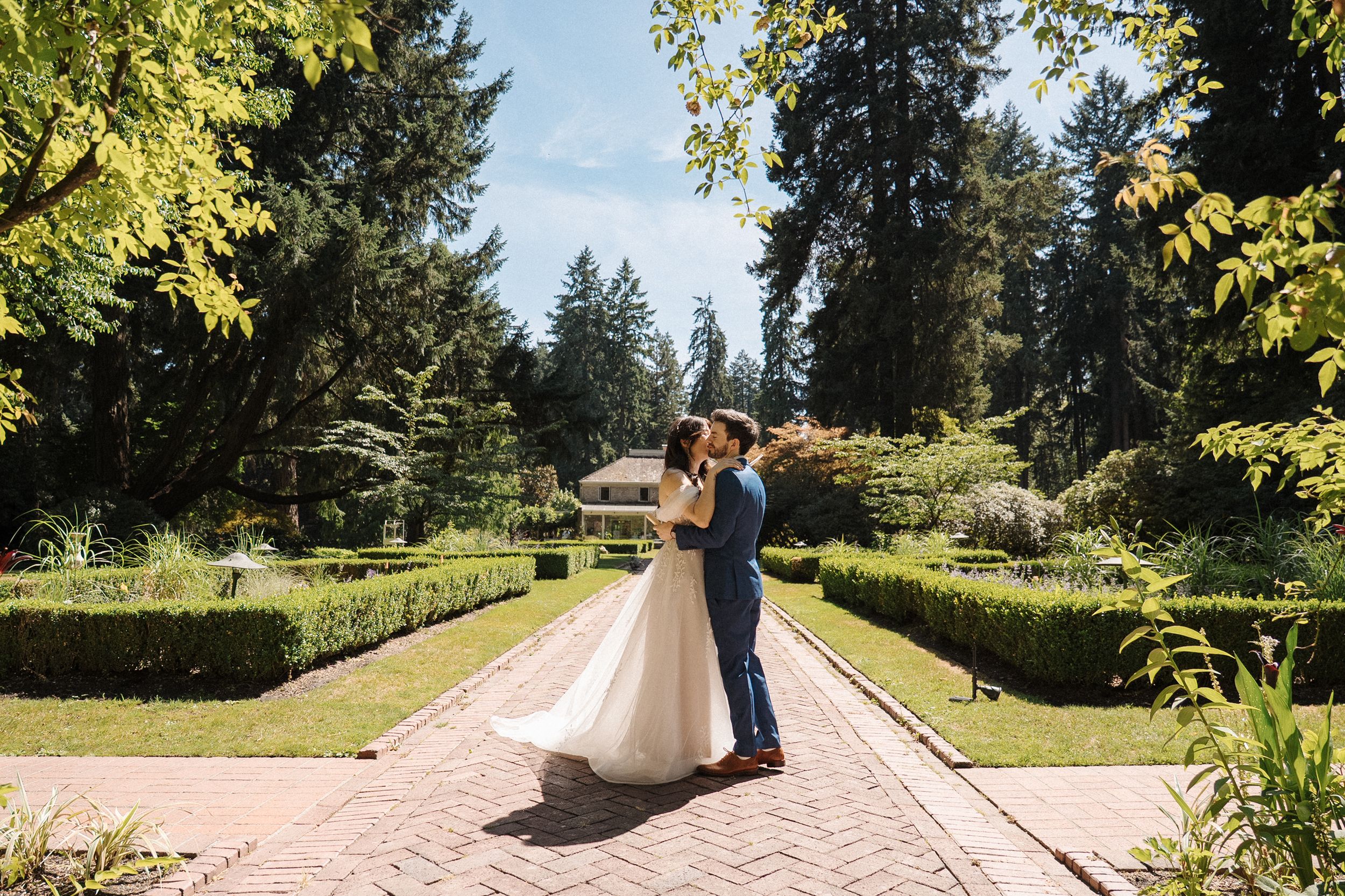 Elegant couple embraces in a sunlit garden with evergreen trees and manicured hedges in the background.