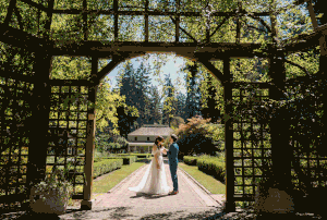 A couple poses under a wooden garden arch surrounded by lush greenery in an intimate outdoor wedding setting.