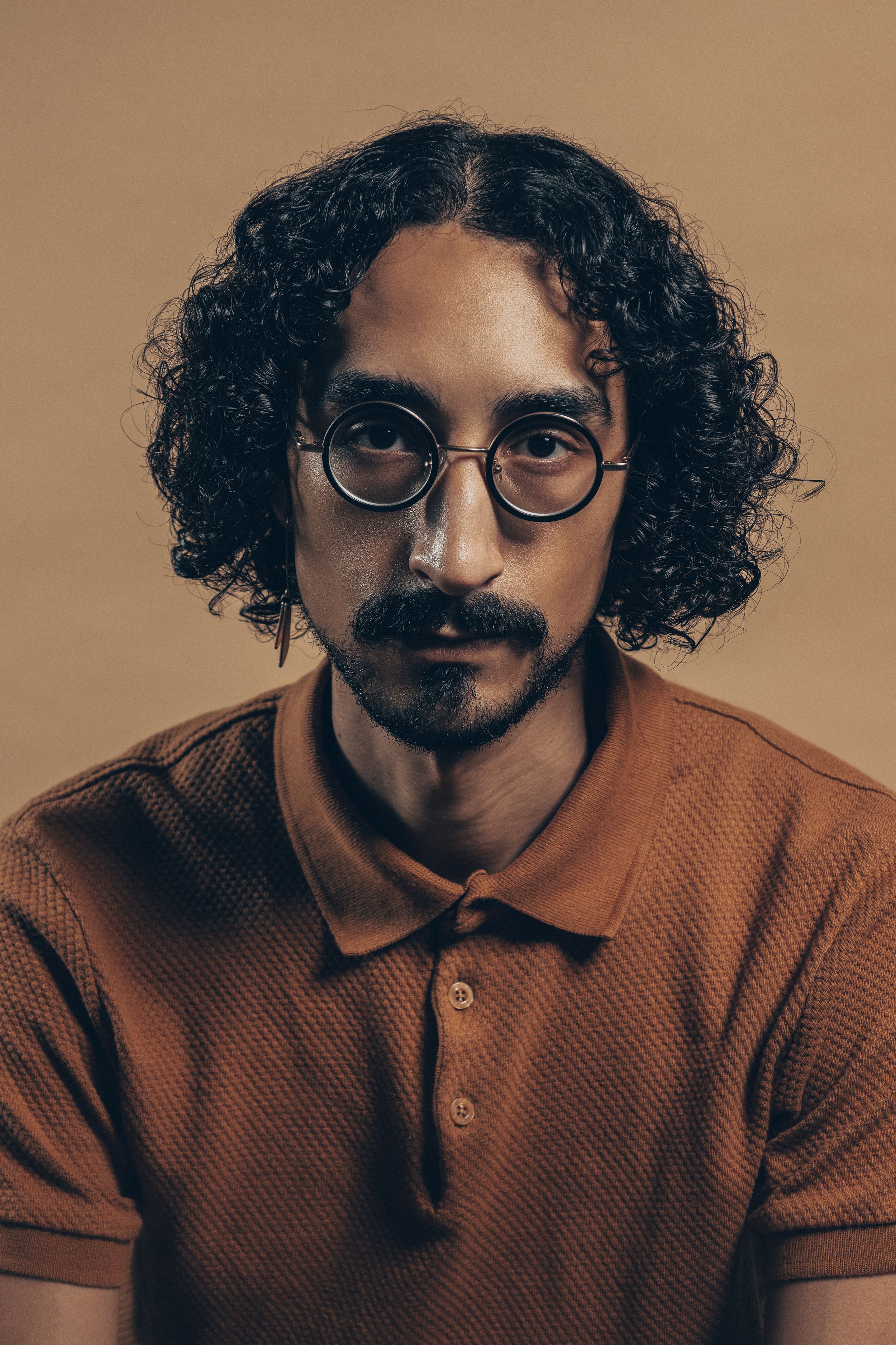 Portrait against brown background of person with curly dark hair and round glasses wearing brown polo shirt.