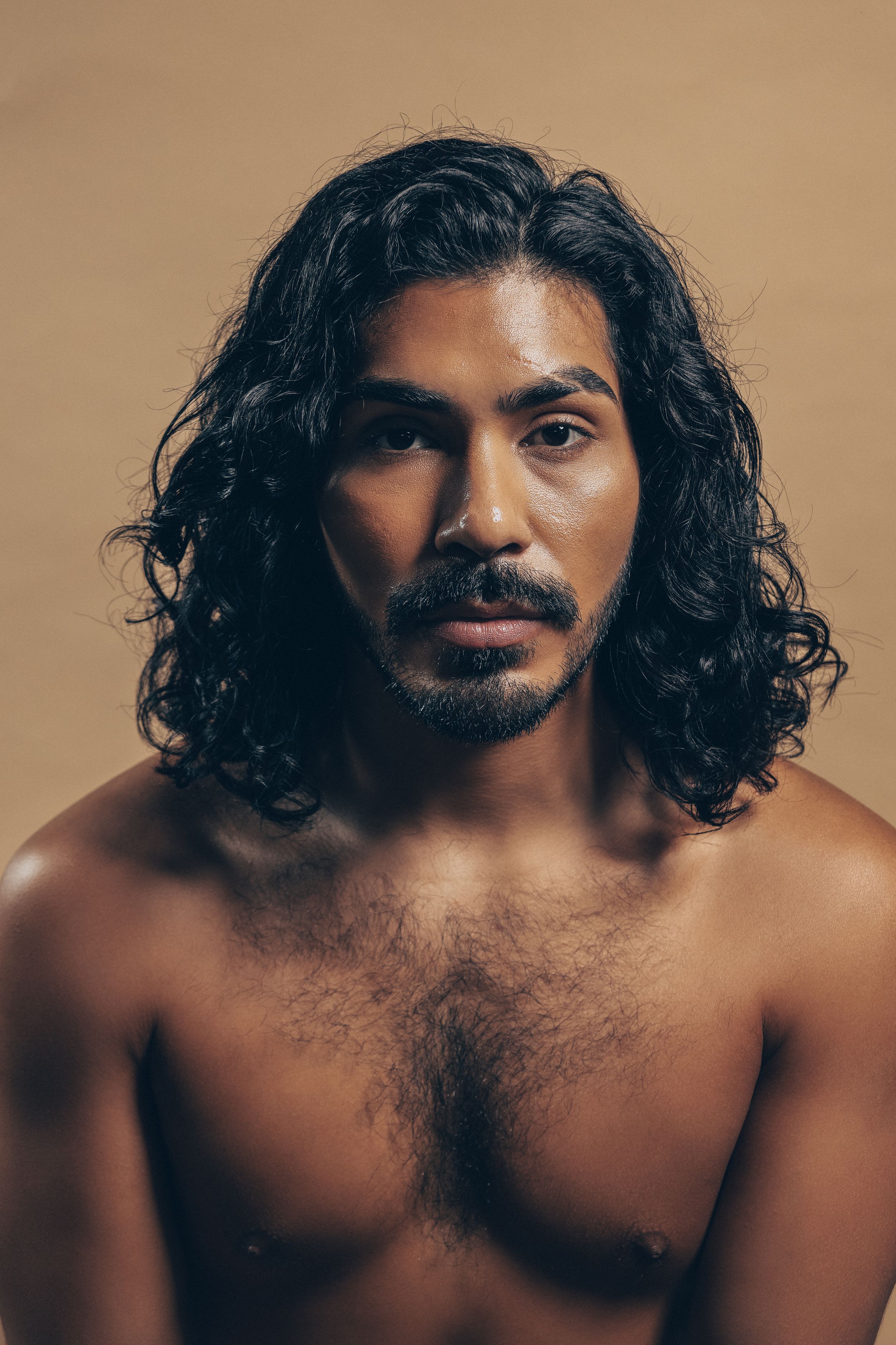 Artistic portrait of a shirtless person with long dark wavy hair and a mustache against a brown background.