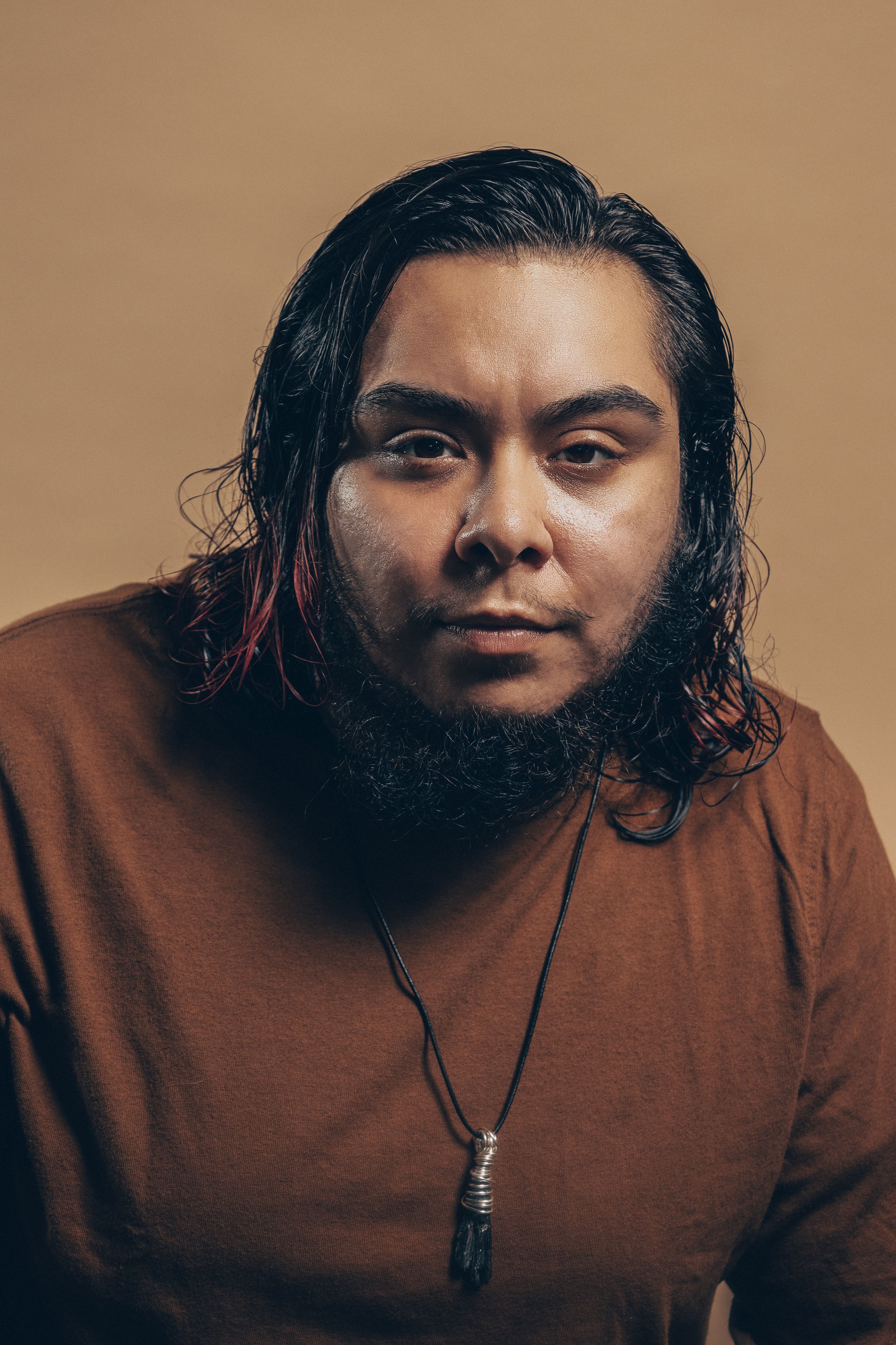Portrait against brown background of person with long dark hair wearing brown shirt and pendant necklace.