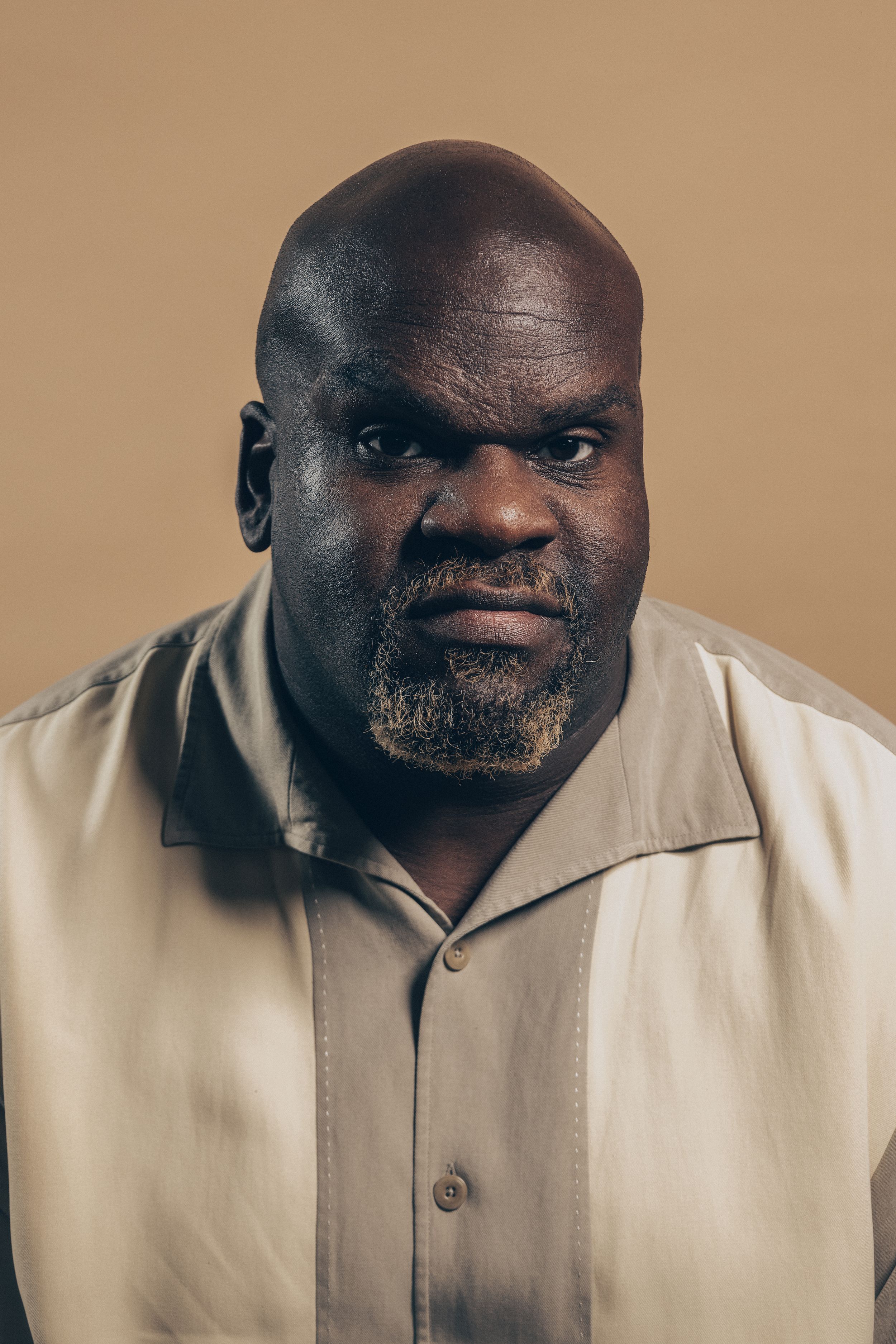 Portrait of person wearing beige button-up shirt against tan background with deep contrast and dramatic lighting.