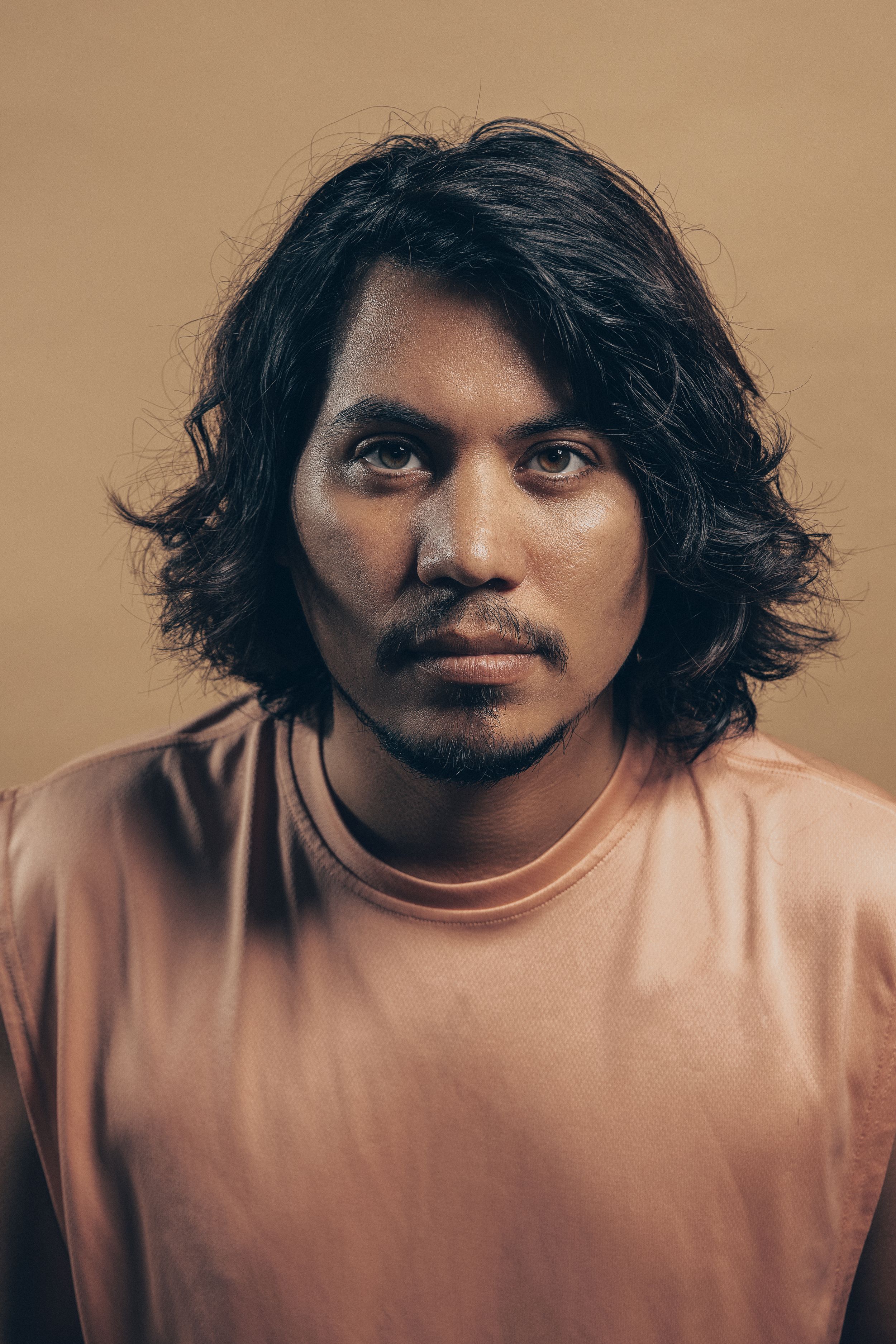 Portrait of person with long dark wavy hair wearing peachy pink sweatshirt against brown background.