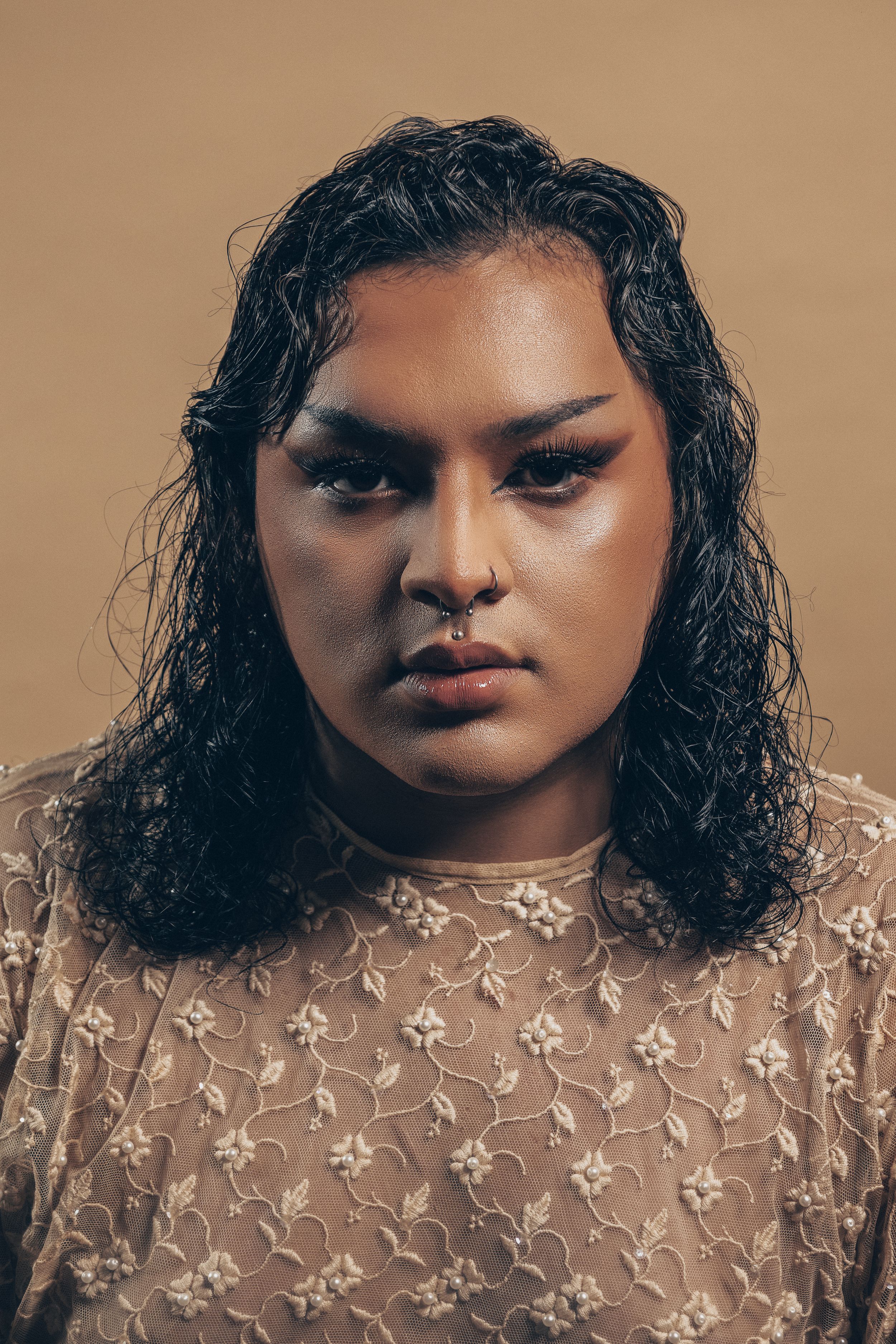 A portrait showcasing wet curly dark hair, makeup, and a sheer floral lace top against a neutral background.