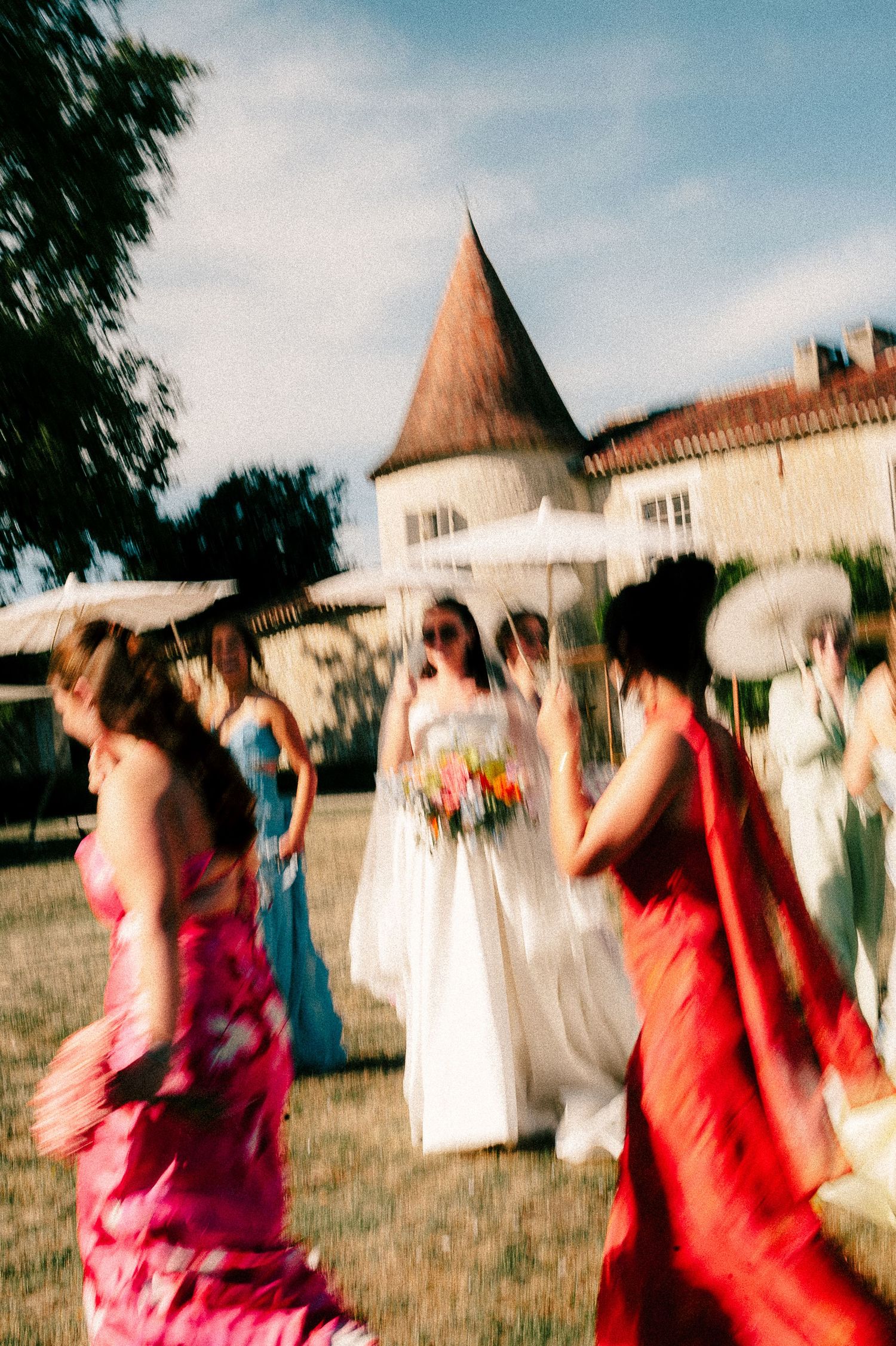 Motion-blurred dancers in colorful dresses spin and twirl in front of a historic building with a tower under blue skies.