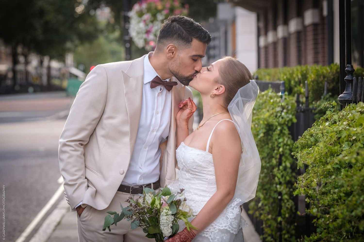 A couple shares a romantic kiss wearing formal attire during their wedding photos against a lush garden backdrop.