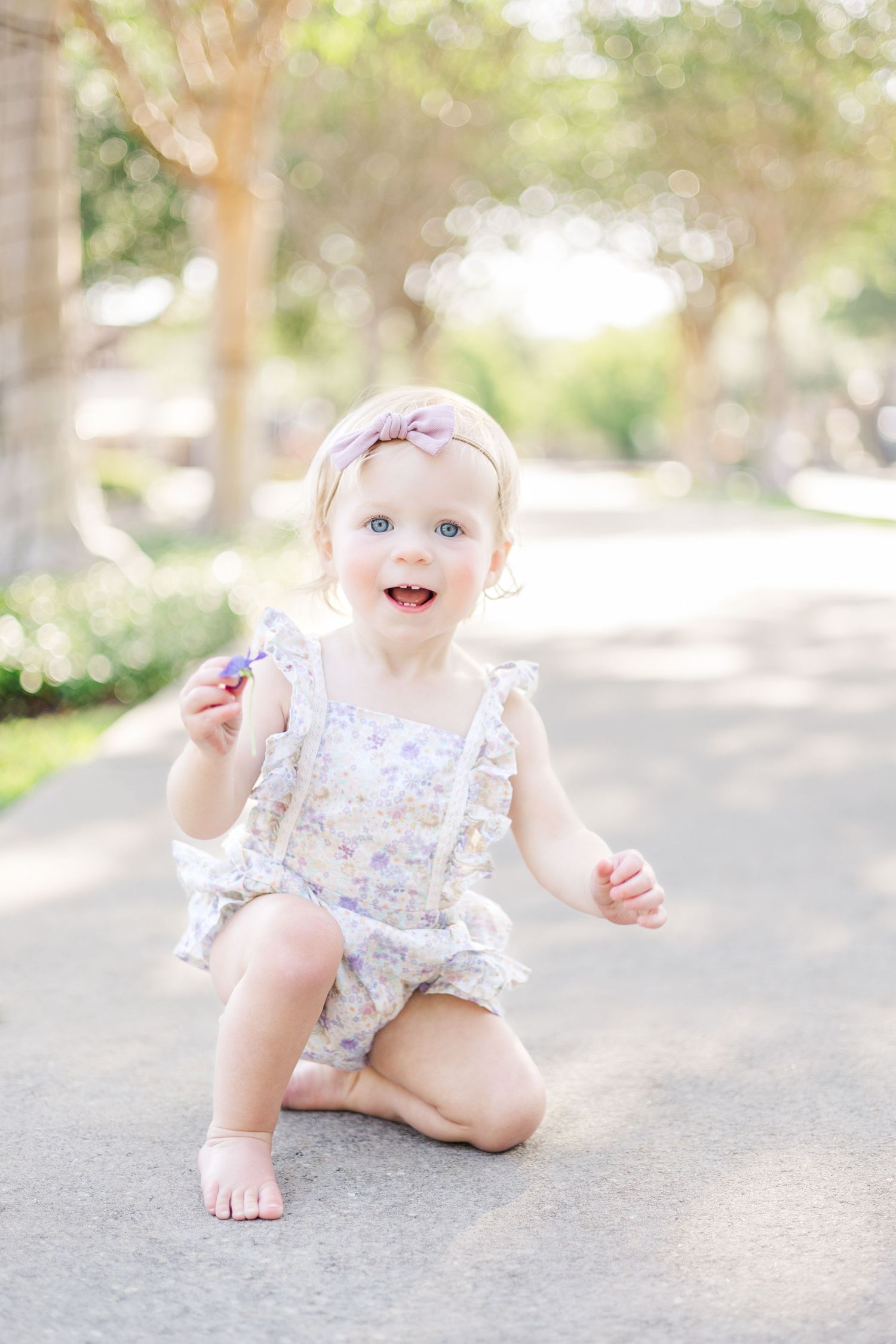 Baby in floral romper crawling on concrete path surrounded by trees.