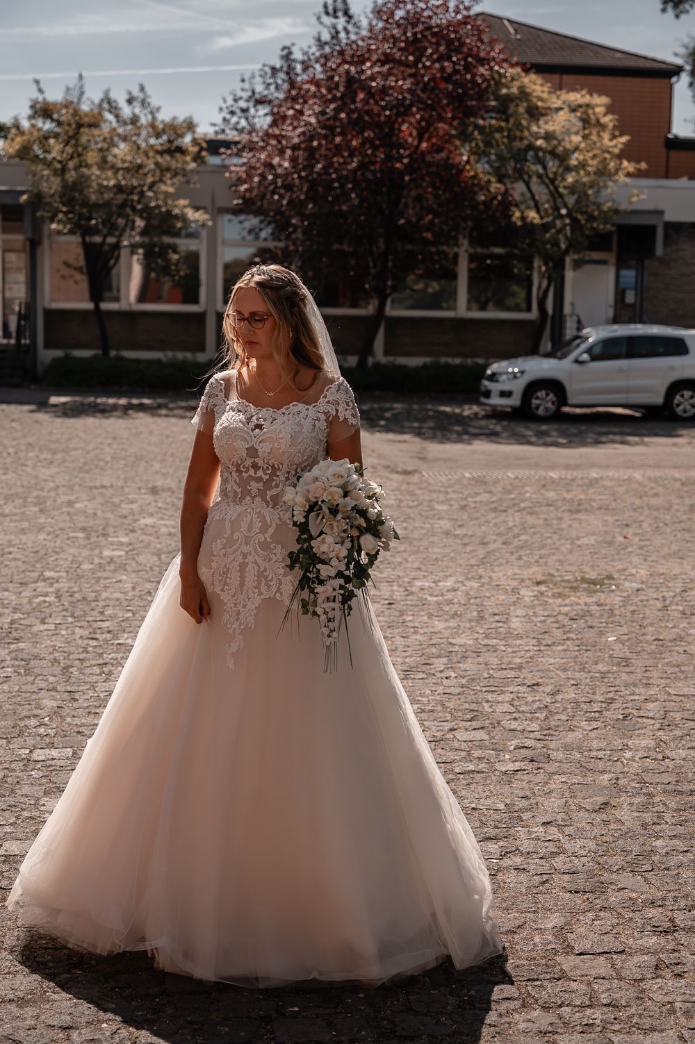 A bride in a flowing ballgown style dress stands in a paved outdoor area near buildings.