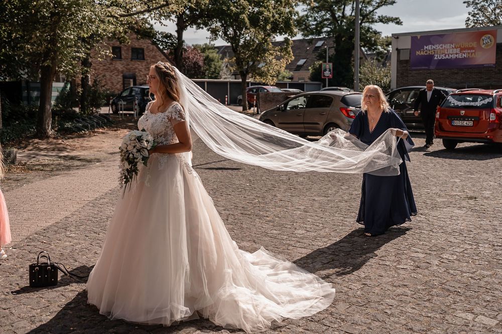 A flowing wedding veil catches the breeze as it trails behind a bride in a tulle gown on a cobblestone street.