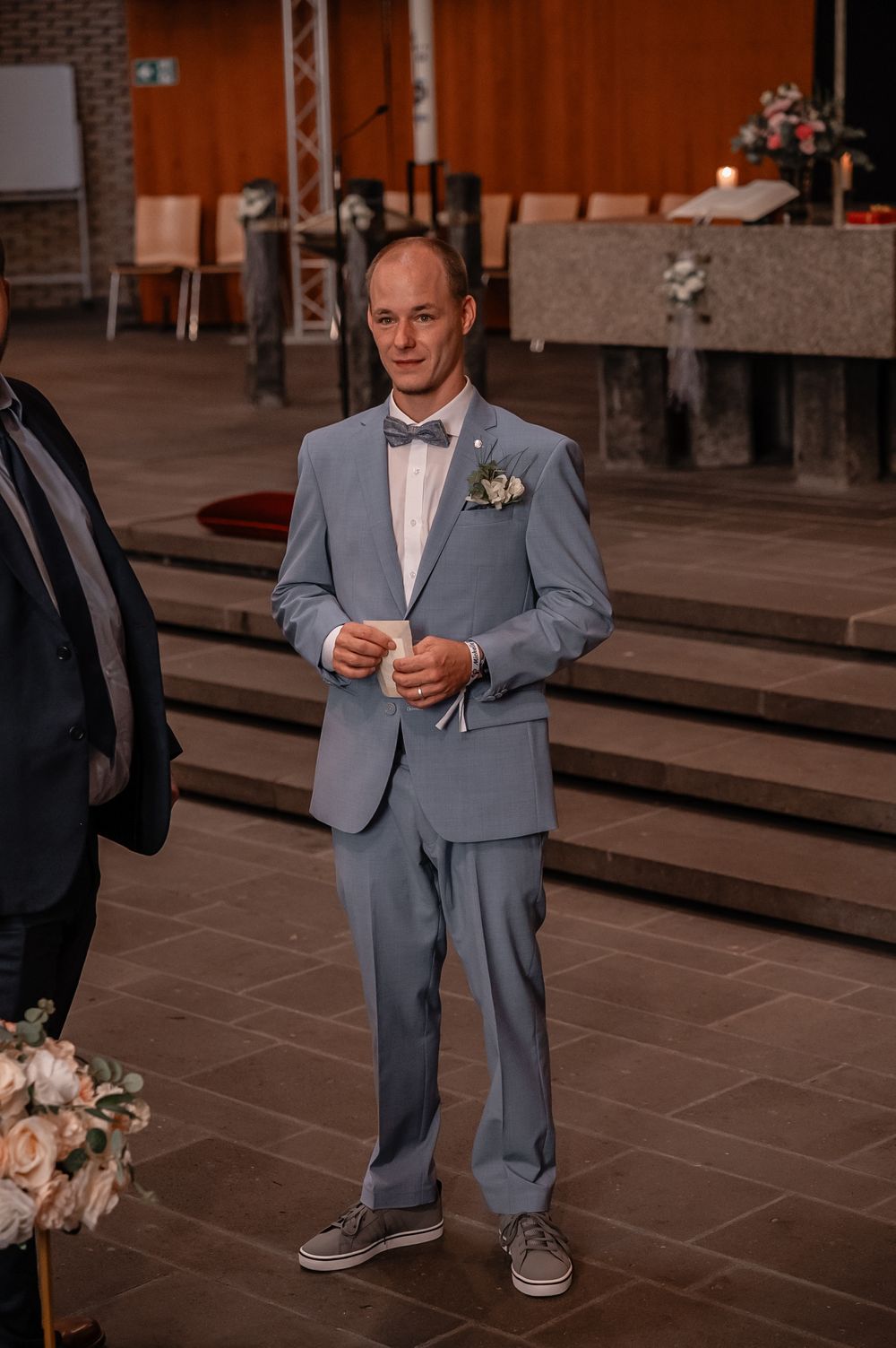 Person in a light blue suit and bow tie standing on stone tiles at an indoor venue.