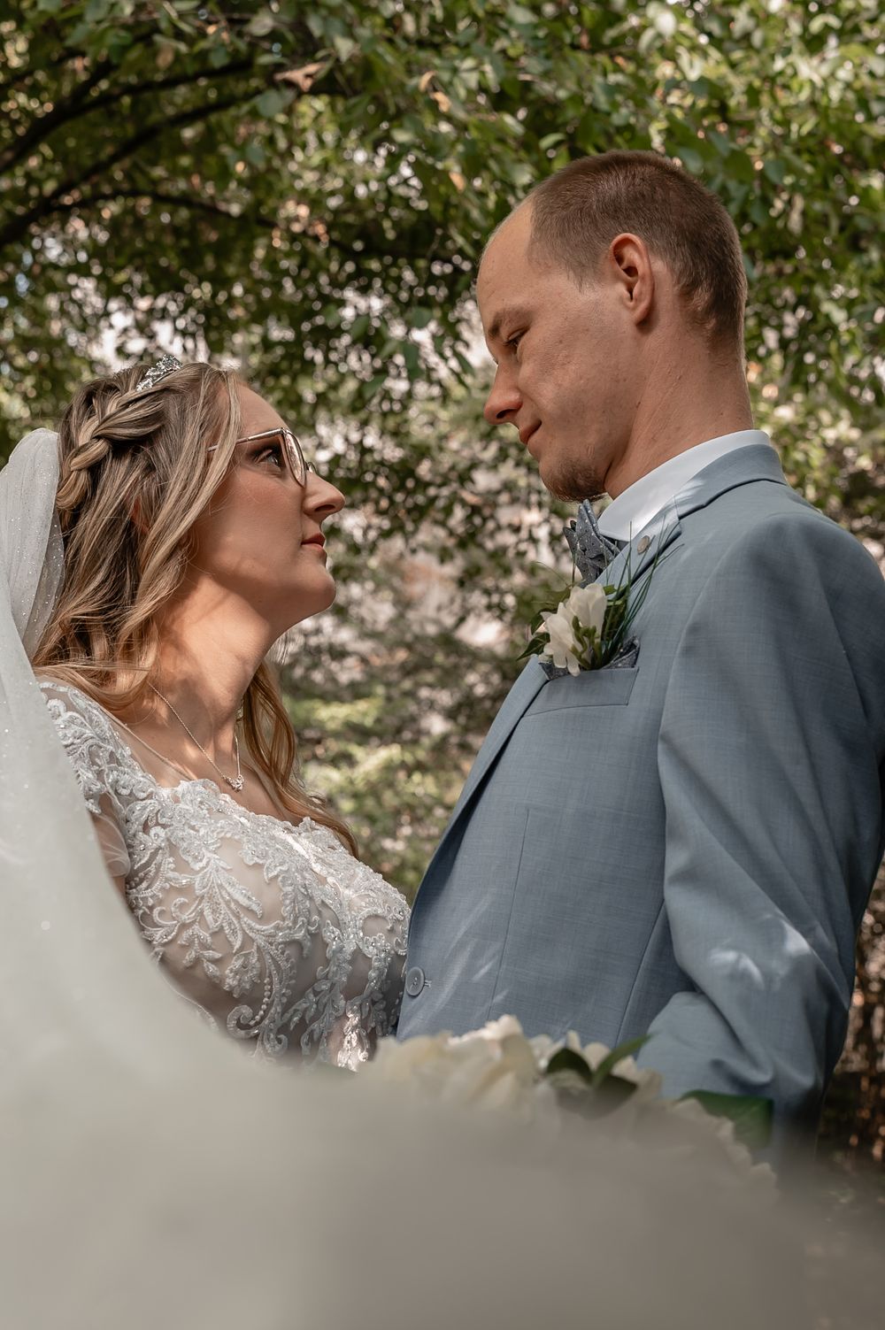 A romantic wedding couple embraces outdoors with the bride wearing a delicate lace dress and veil.