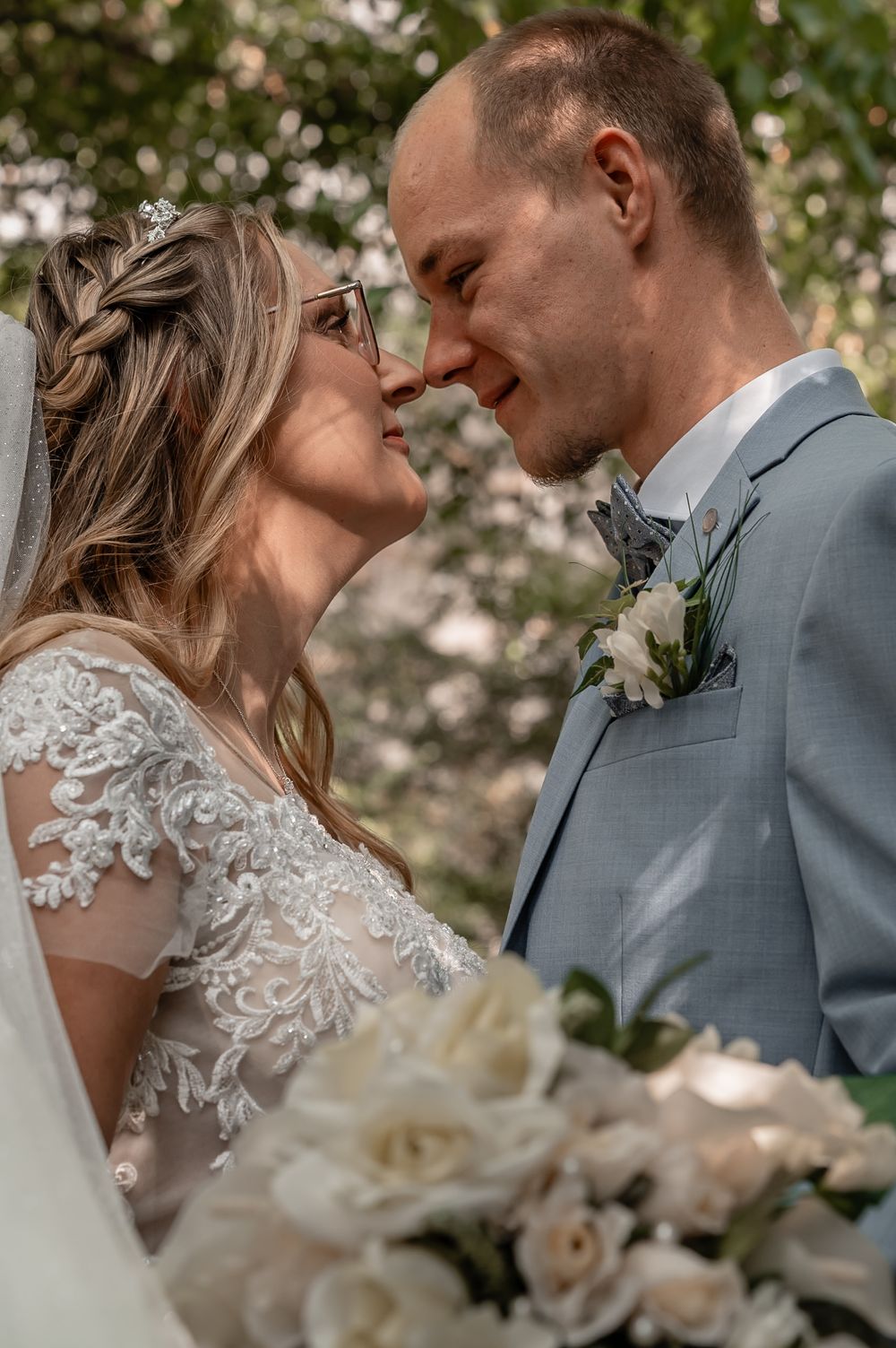 Romantic wedding couple shares intimate moment under flowering trees with bride wearing lace detailed dress.