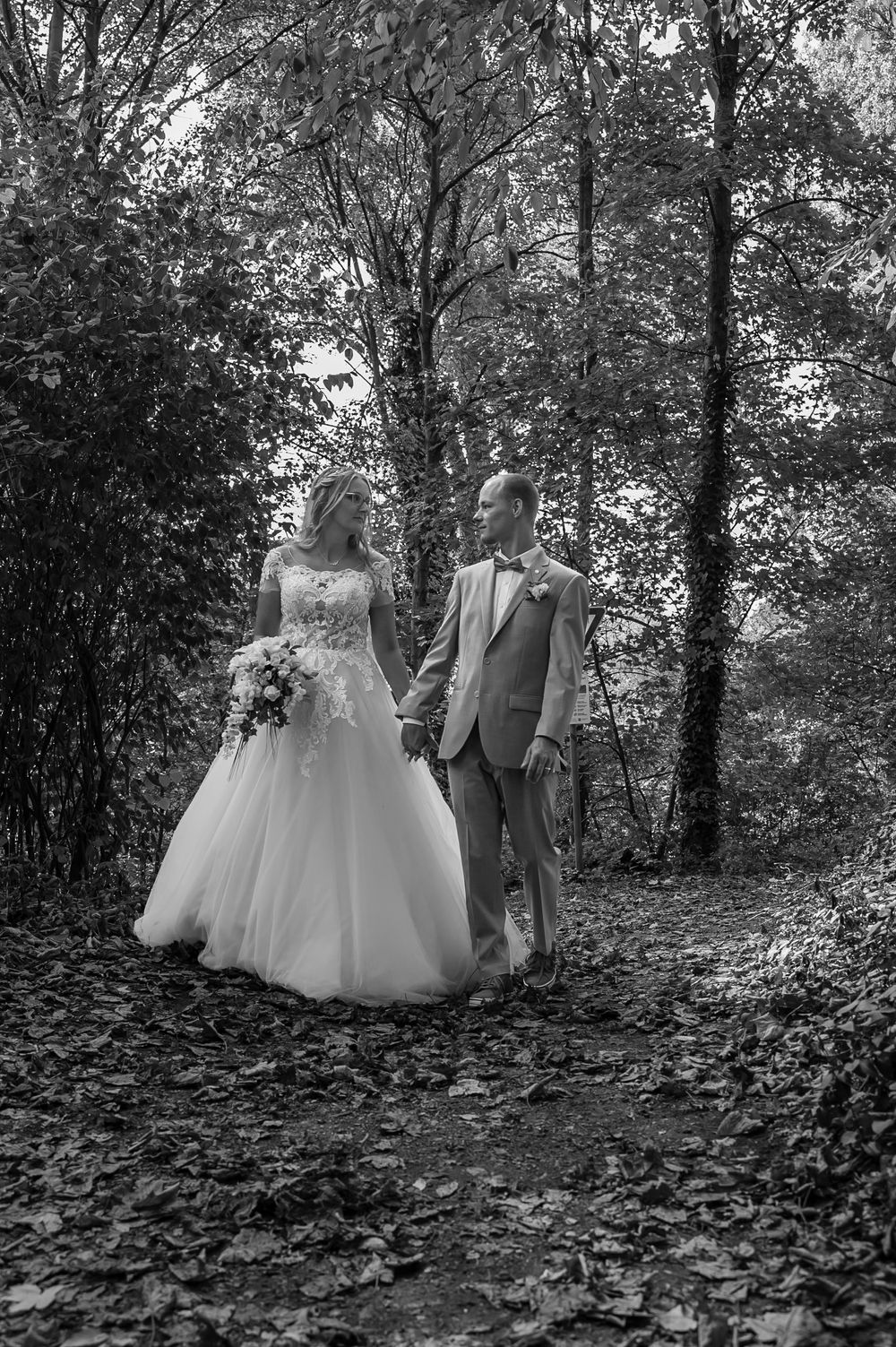 Black and white wedding portrait of couple standing among fallen autumn leaves in wooded area.