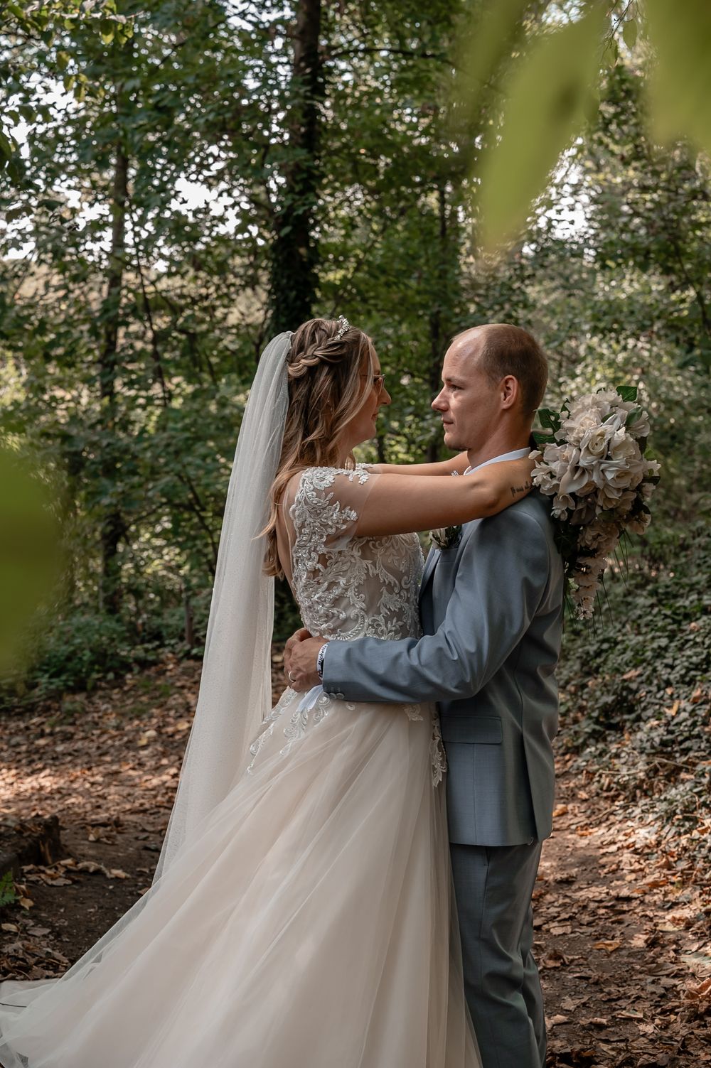 A couple shares an intimate moment in a wooded area, with the bride wearing a flowing white dress and groom in grey suit.