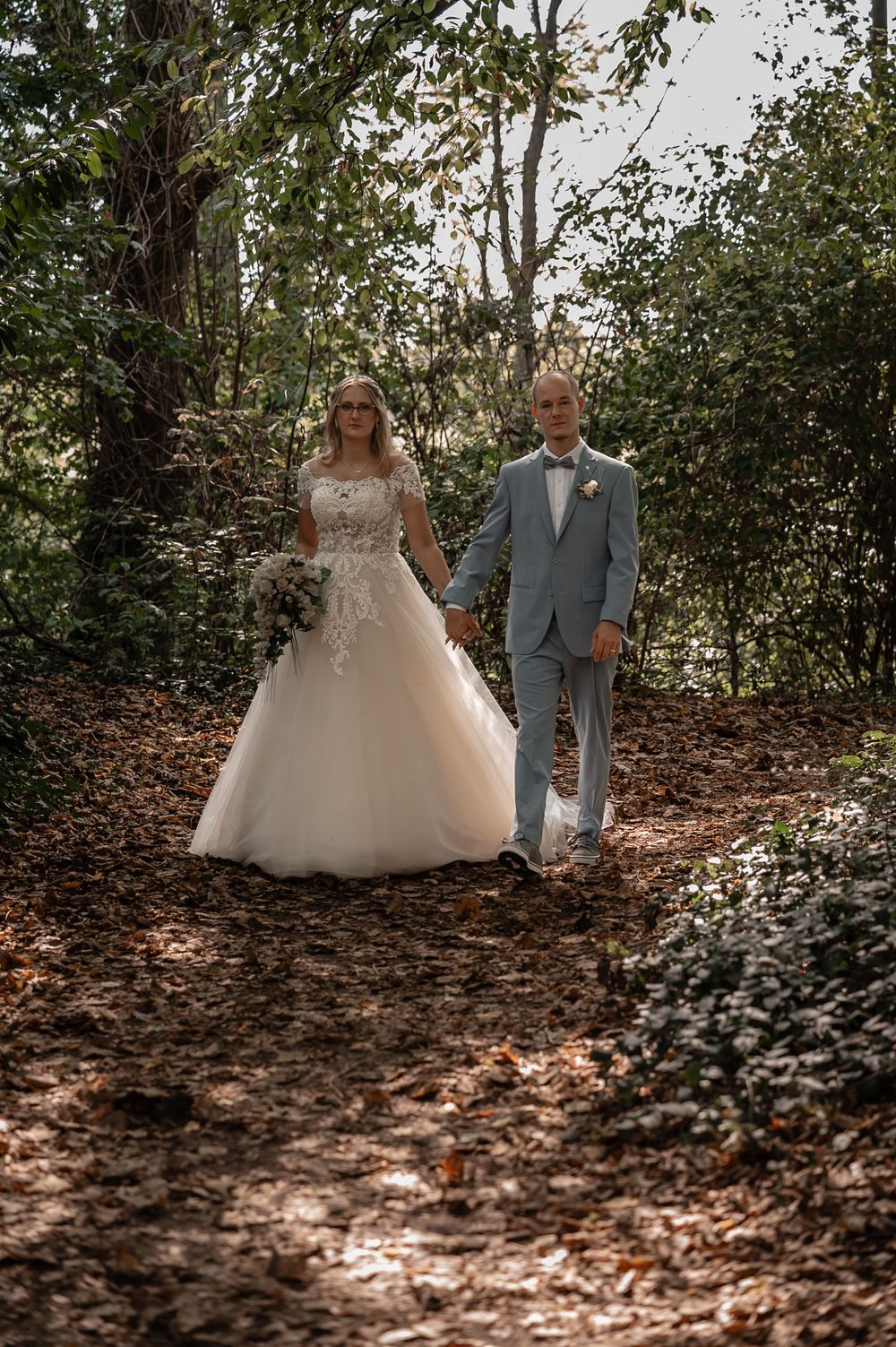 Wedding couple walking through a wooded path with dappled sunlight filtering through the trees.