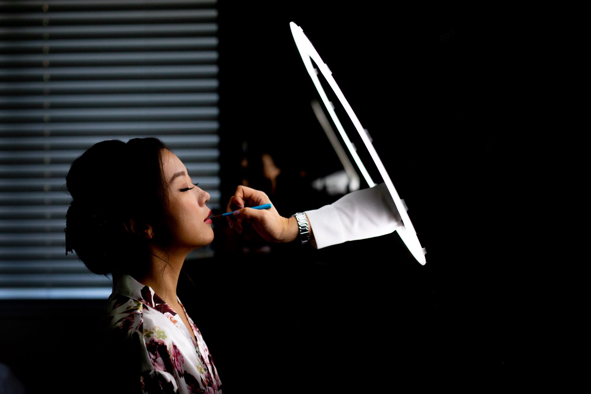 A person in profile view exhales vapor from an e-cigarette against a dark background with horizontal blinds.