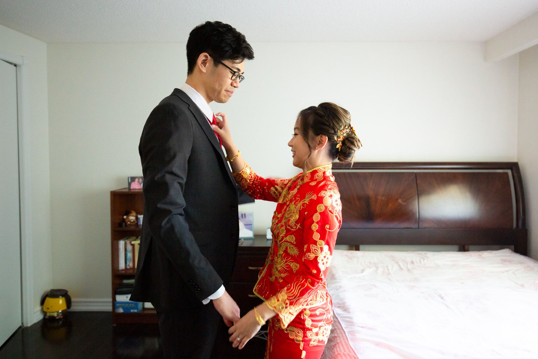 A couple in traditional wedding attire, with the bride wearing a red qipao dress and groom in black suit in bedroom.