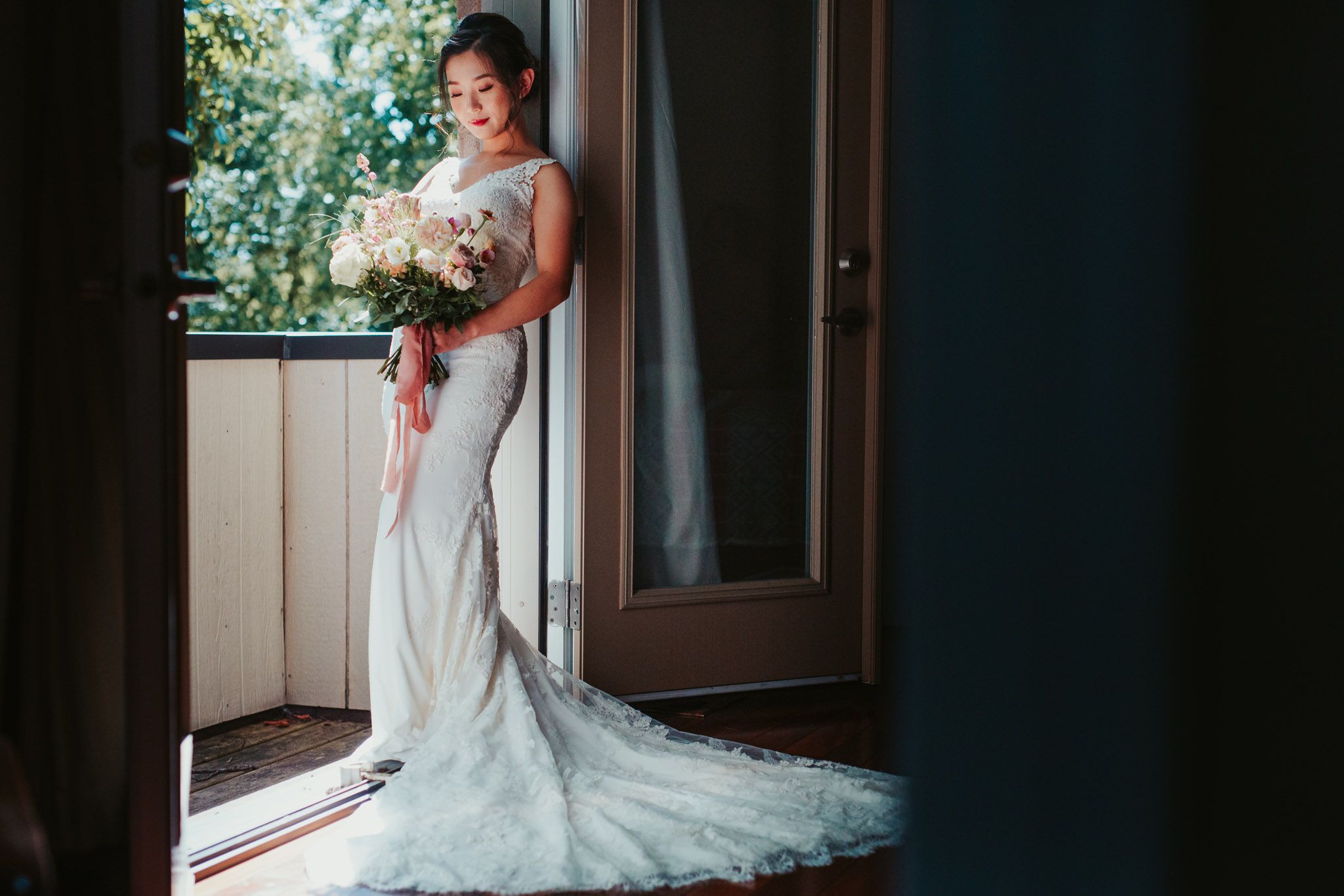 A bride in a fitted lace wedding gown with a long train stands by a window holding a white floral bouquet.