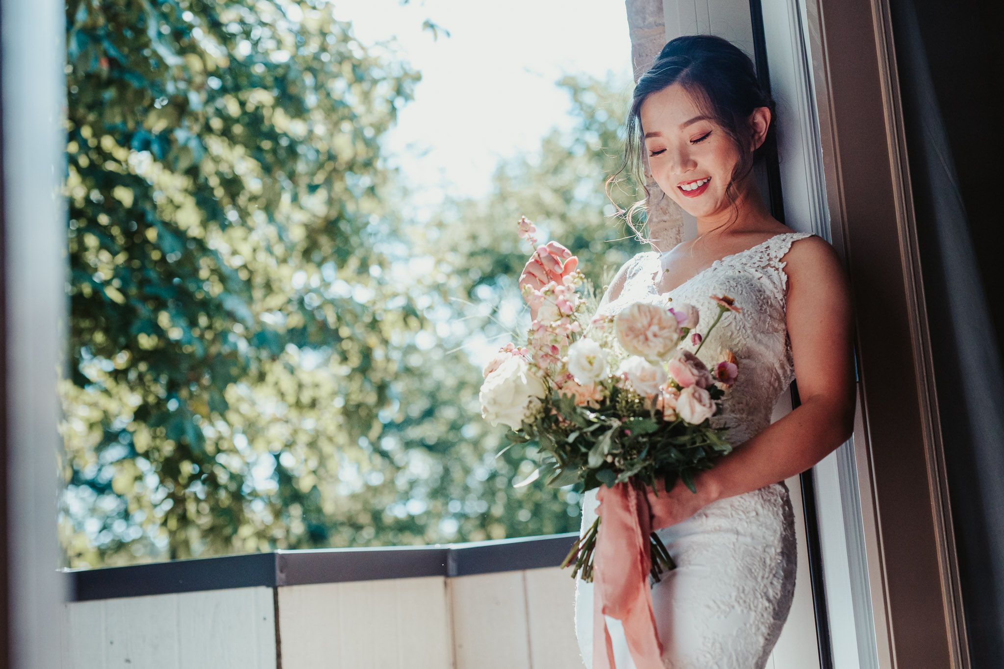 A bride in an elegant white dress smiles while holding a beautiful bouquet near a sunlit window.