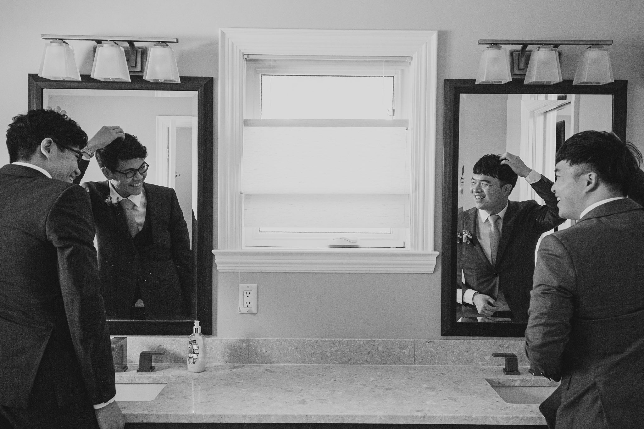A black and white photo of two people looking in mirrors while getting ready in a bathroom.