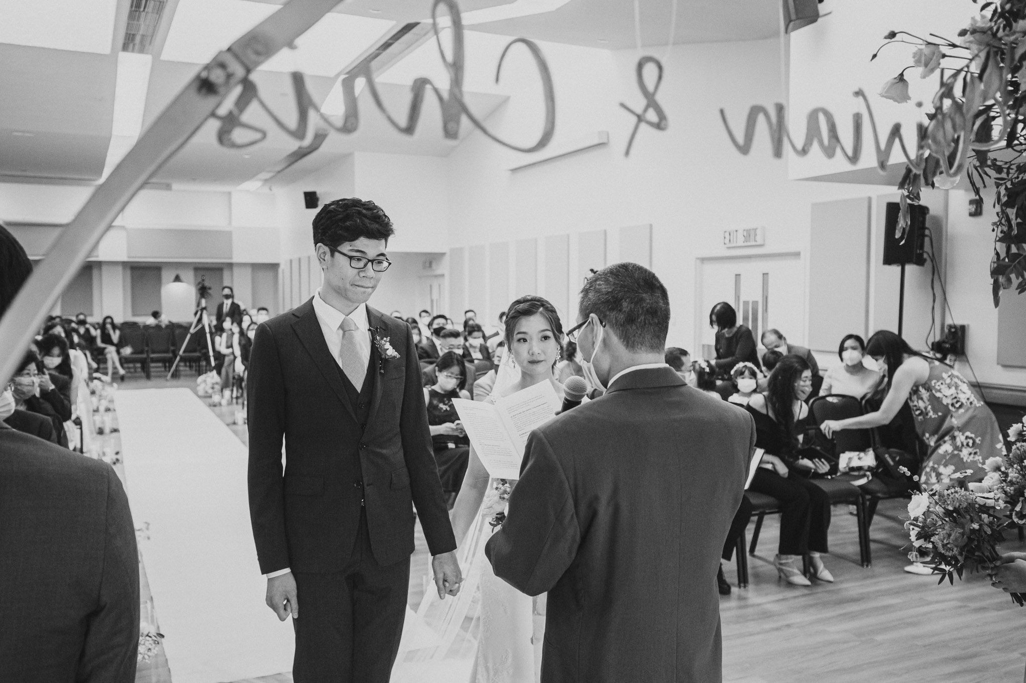 A black and white photo of a wedding ceremony taking place in a reception hall with guests seated.
