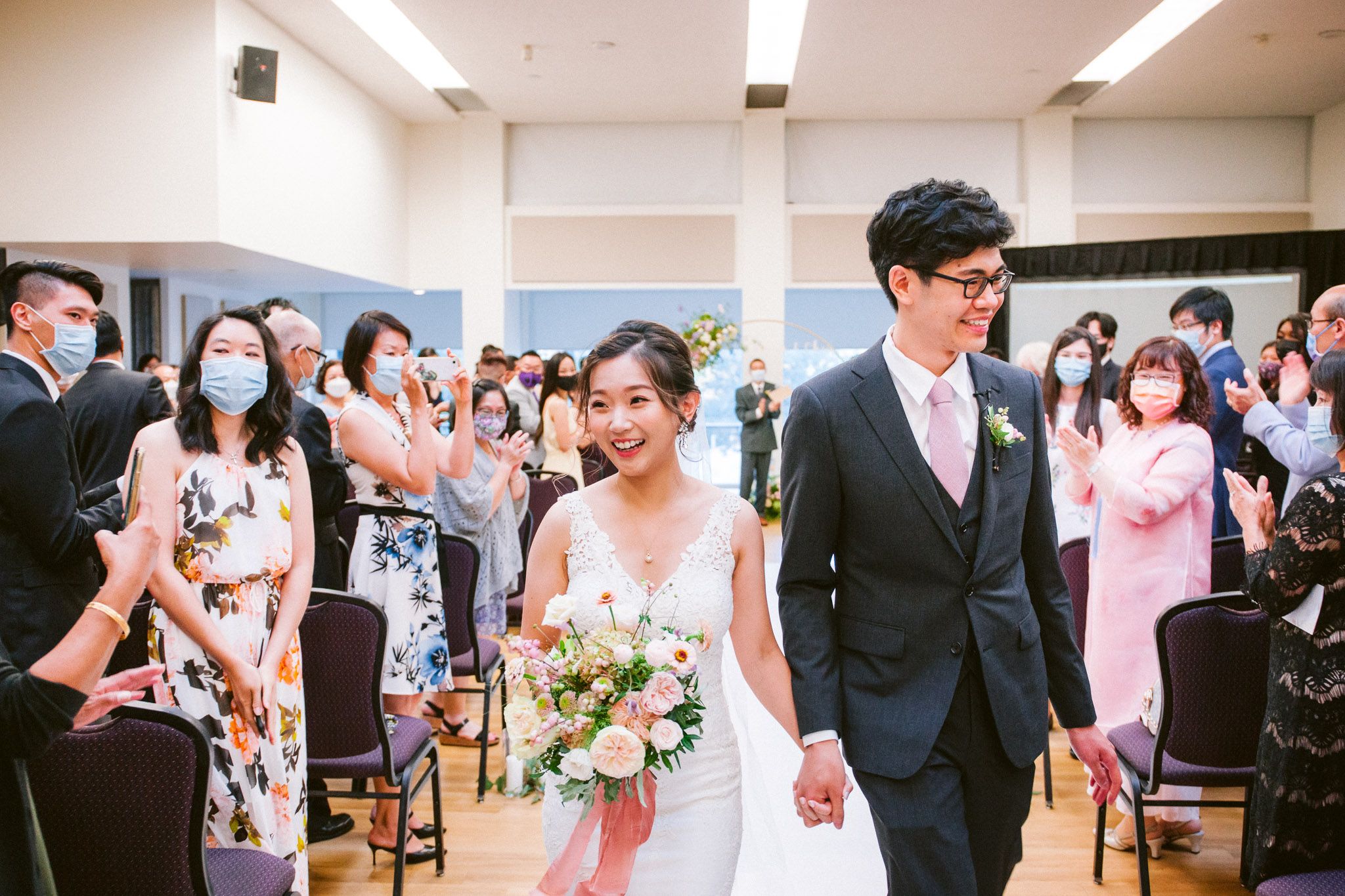 A couple walks down the aisle at their wedding ceremony while guests in masks look on and take photos.