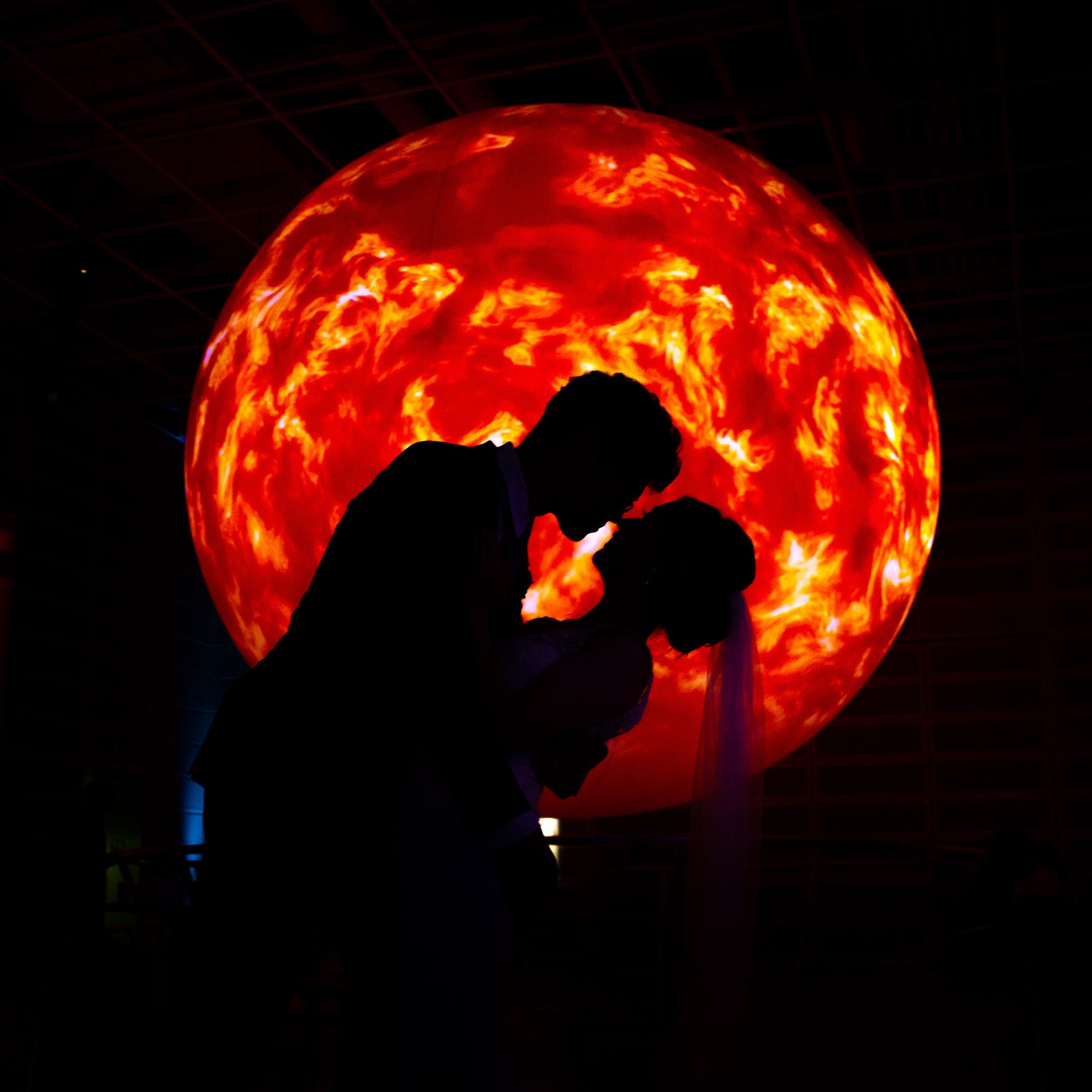 Wedding couple poses under a glowing red arch creating dramatic silhouettes against the dark background.