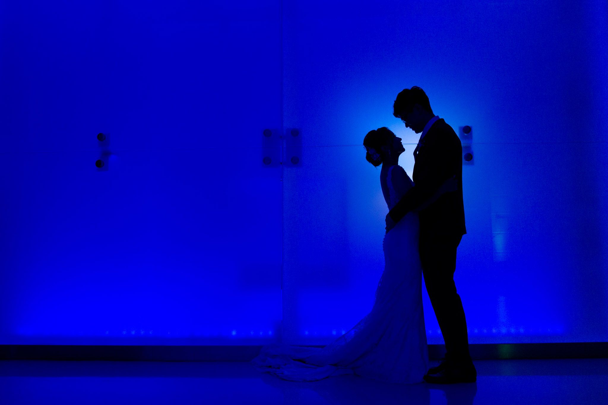 A silhouetted couple embraces against a dramatic blue-lit wall during their wedding reception.