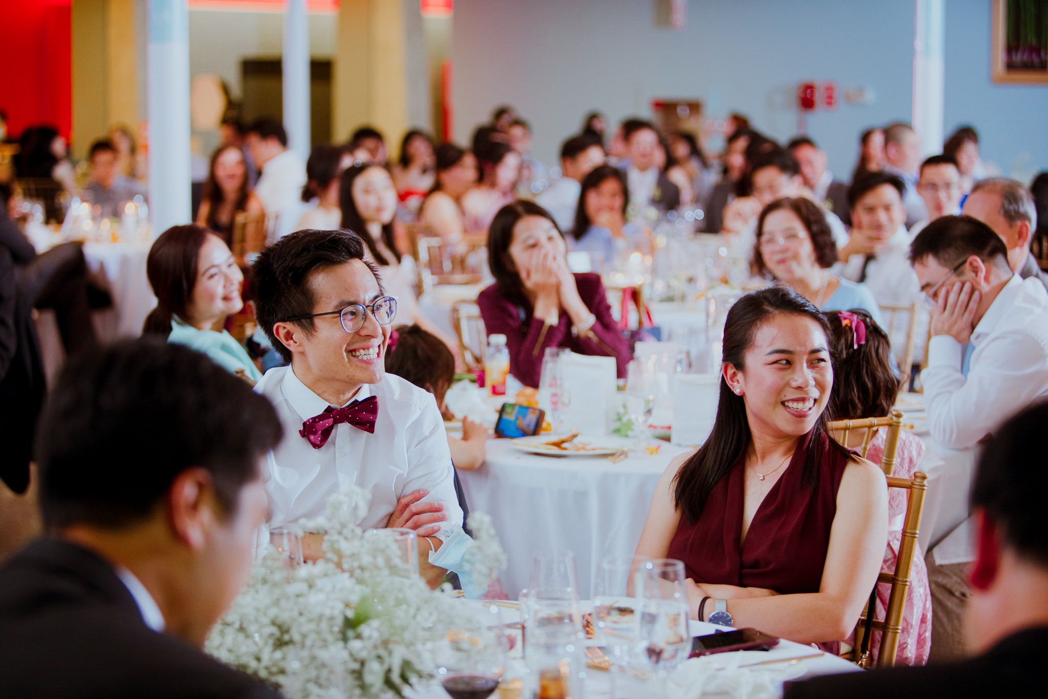 Wedding reception guests seated at round tables with white tablecloths enjoy conversation during an indoor celebration.