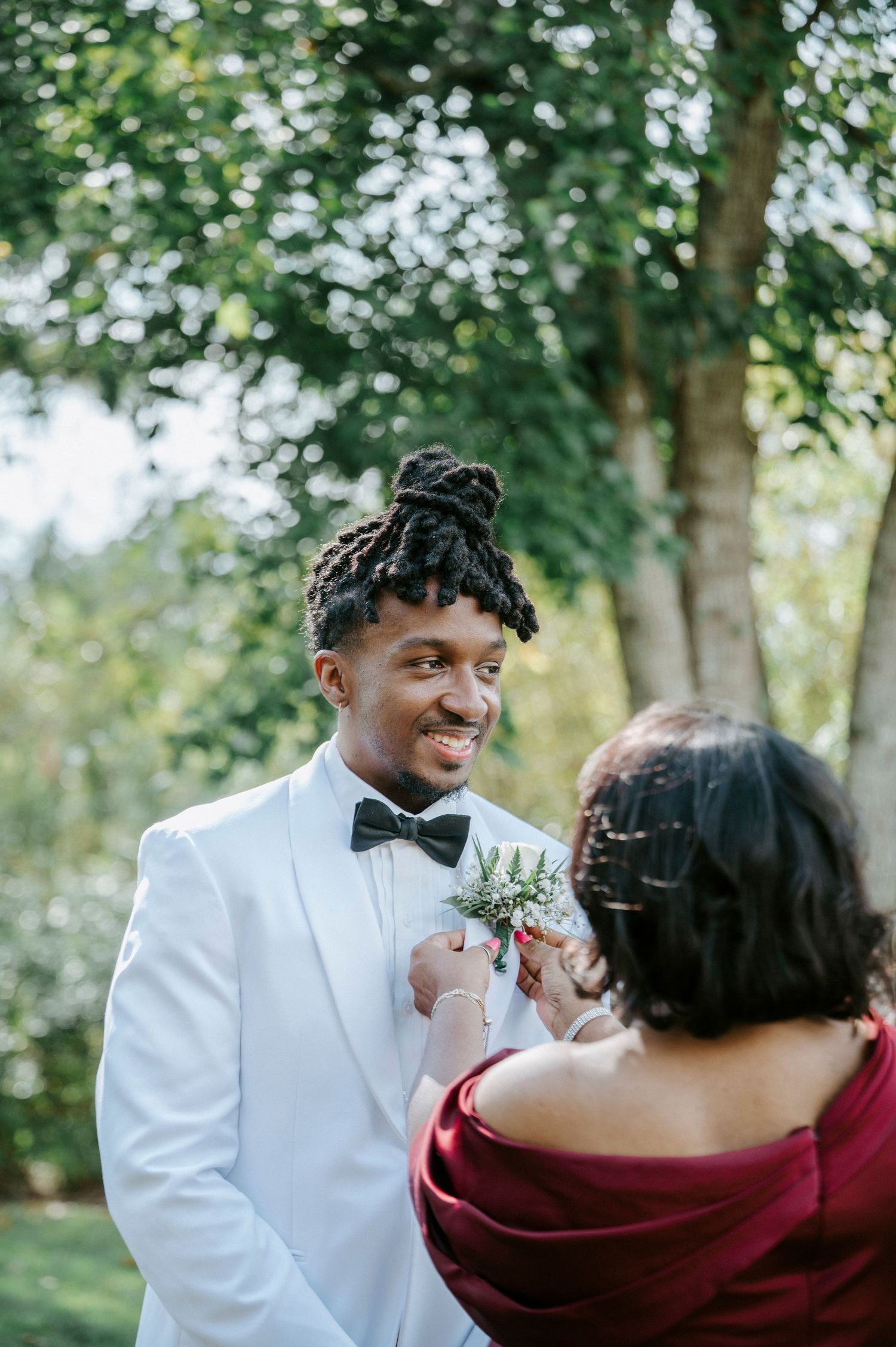 A couple in wedding attire shares an intimate moment outdoors, with the groom in white and bride in burgundy.