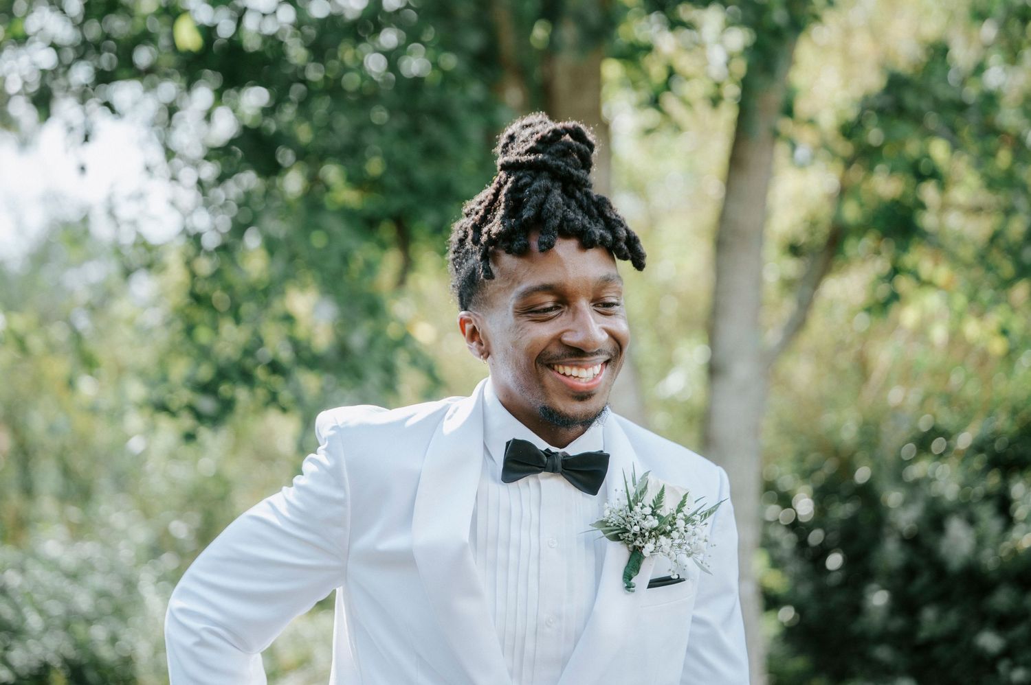 A groom in a white tuxedo jacket and black bow tie smiles joyfully during a wedding celebration.