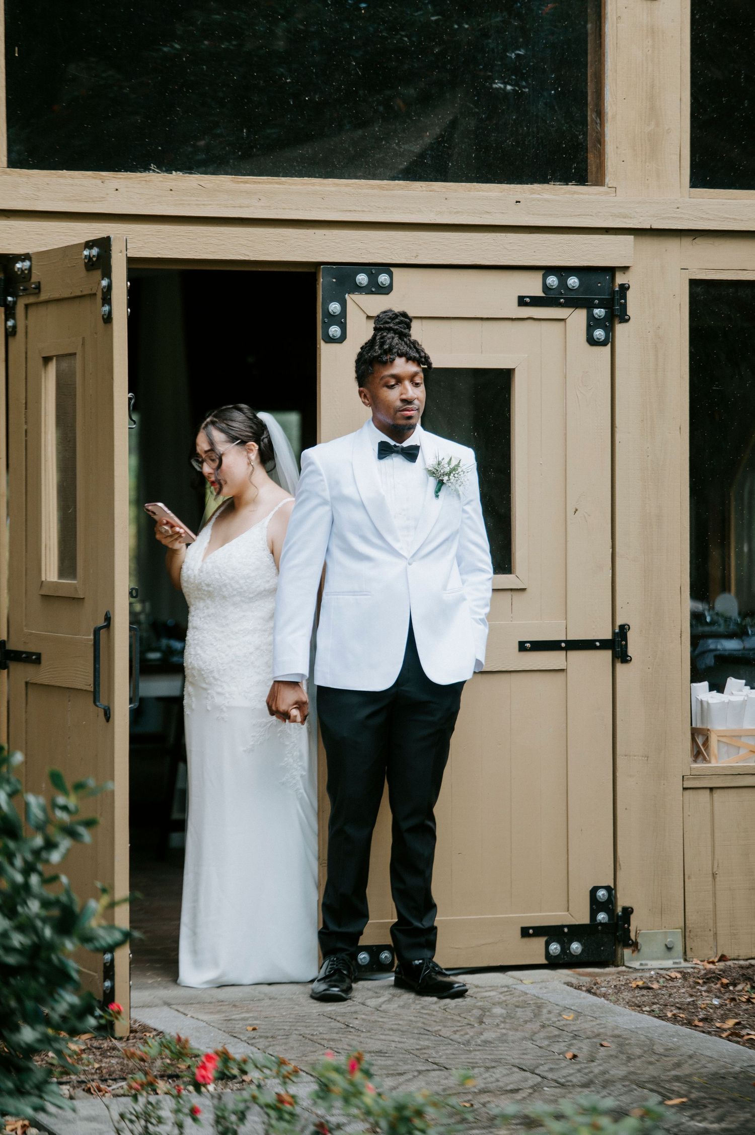 A bride and groom share a playful moment around a corner of a beige building during their first look wedding photos.