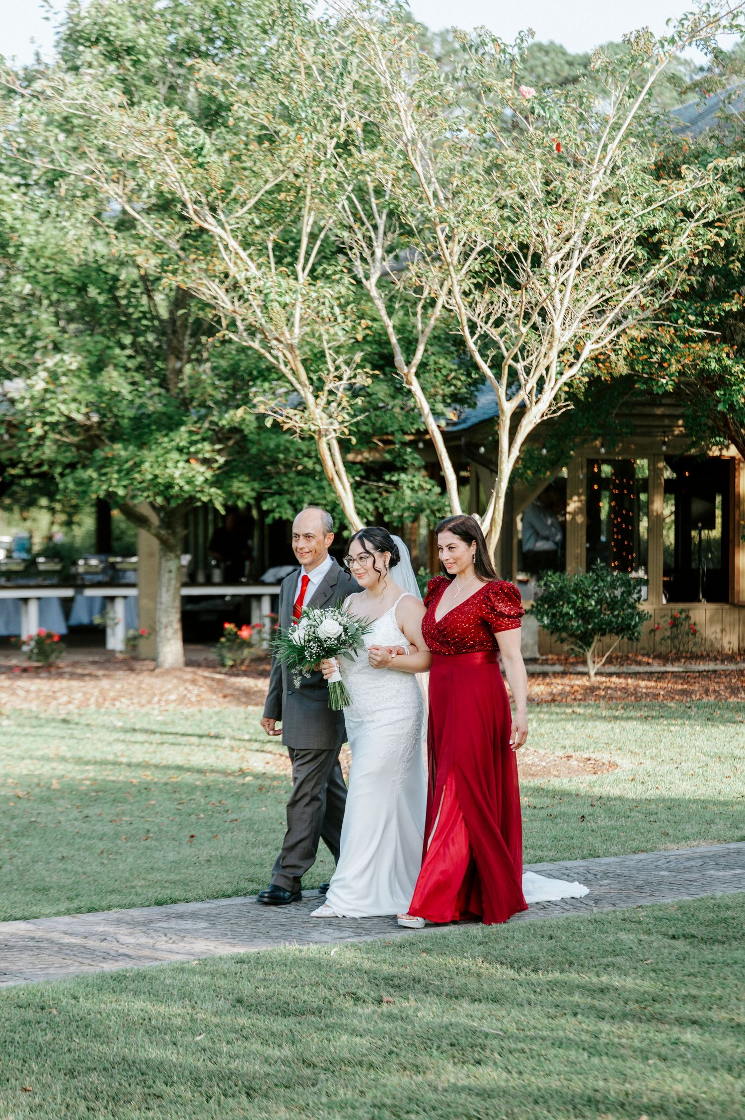 Wedding party in formal attire poses playfully on a grassy lawn surrounded by trees during an outdoor celebration.