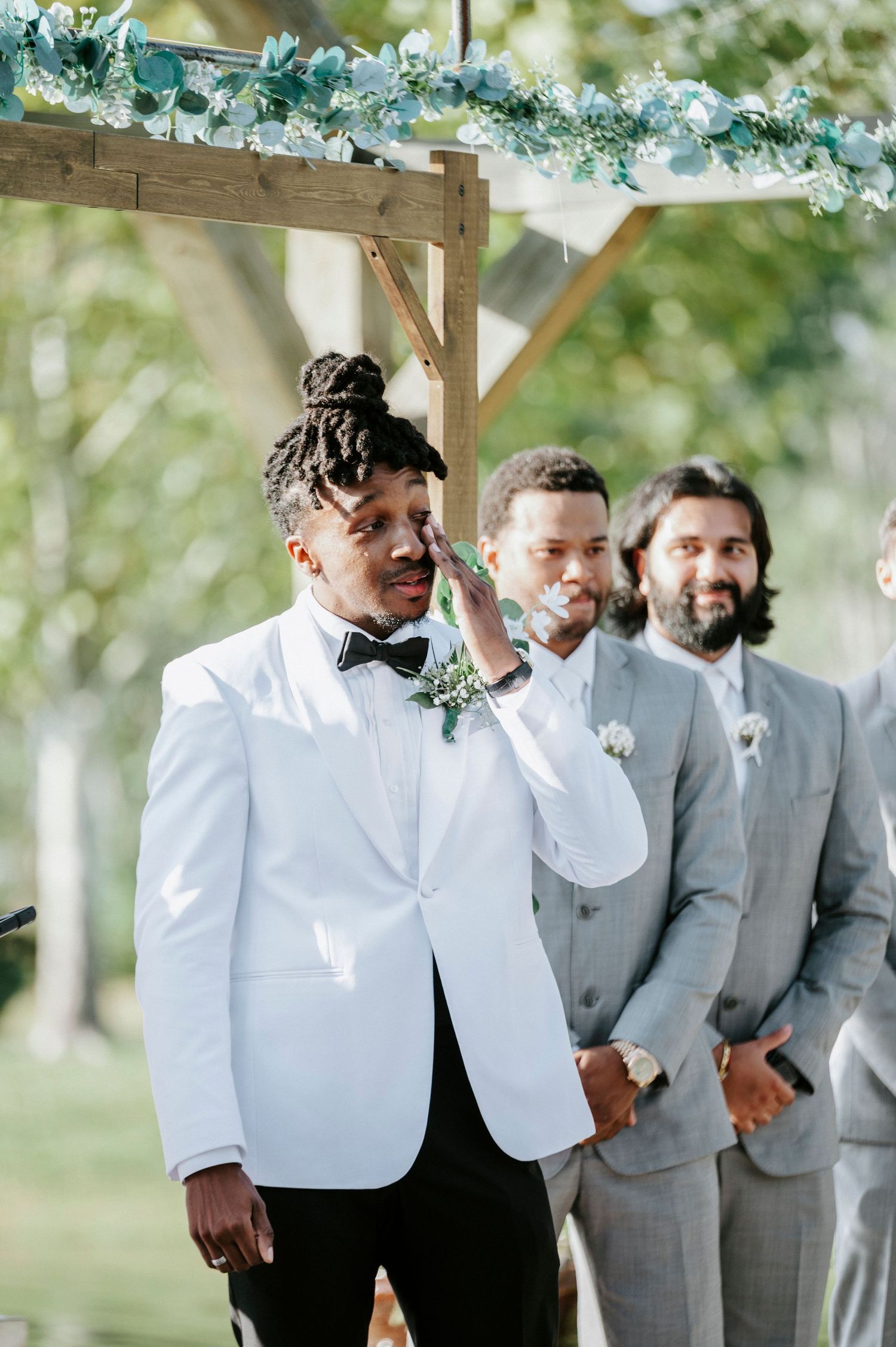 Wedding party members in gray suits and white attire share a candid moment during an outdoor ceremony.