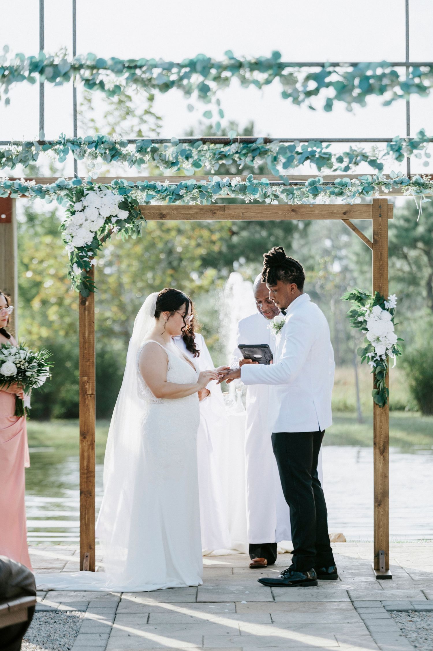 A wedding ceremony takes place on a waterfront patio with white floral decorations and wooden arch.