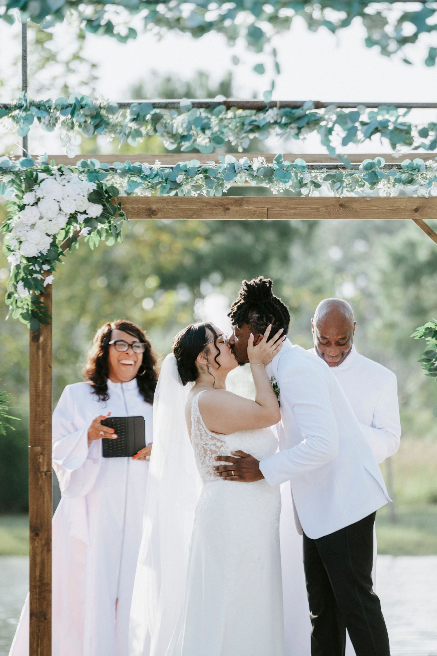Wedding party wearing white outfits embraces in a joyful group hug under a wooden pergola outdoors.
