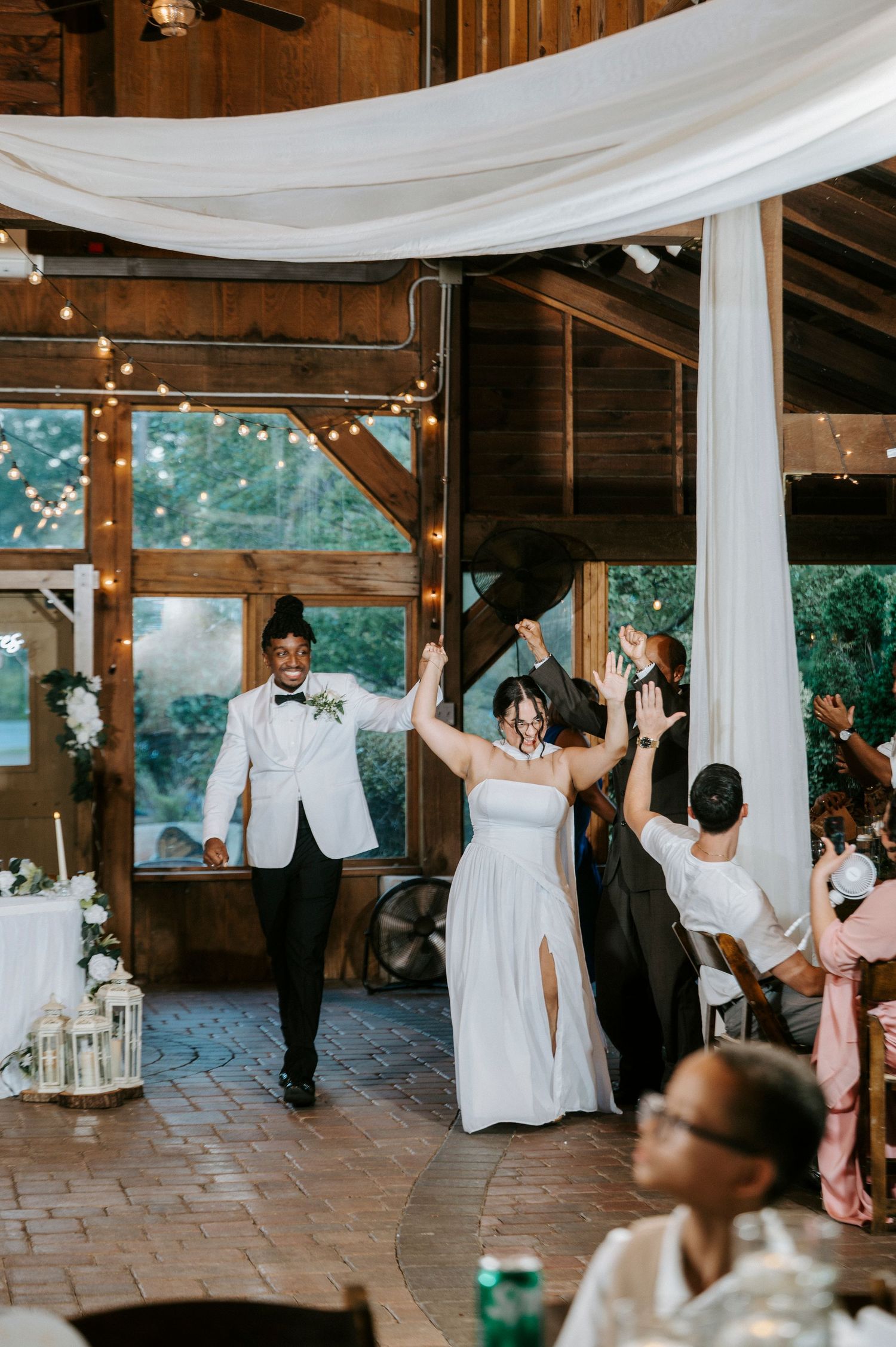 Wedding guests dancing and celebrating at a rustic barn reception with white drapes and string lights.